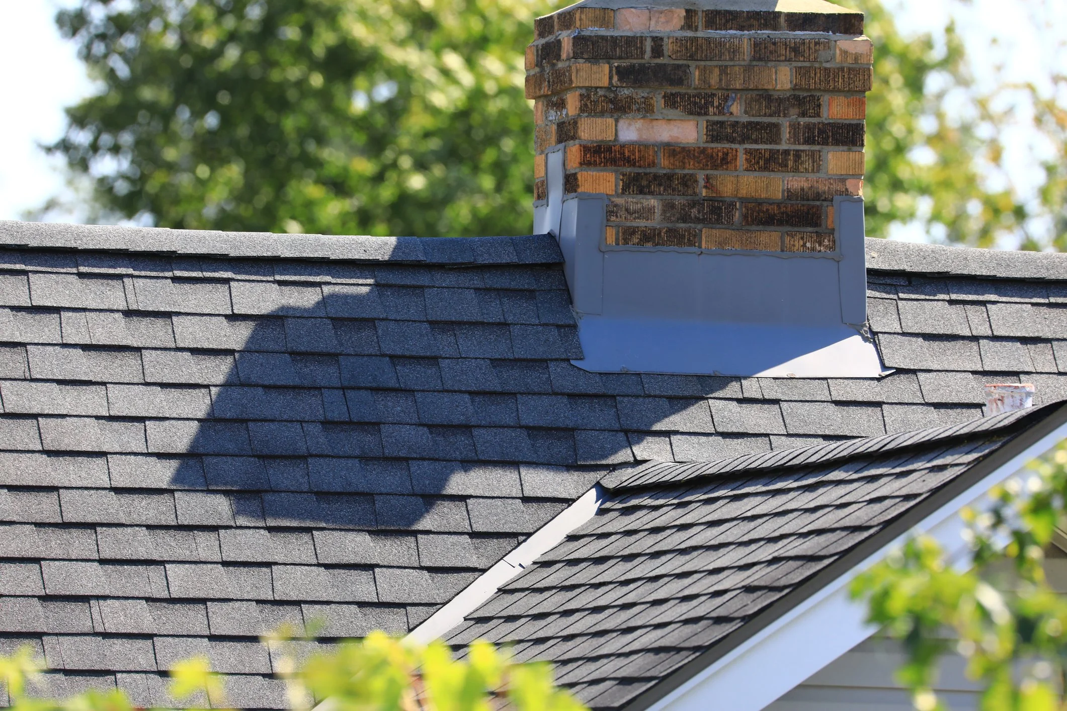 Close-up of a house roof with dark gray asphalt shingles and a red brick chimney, sunlight casting shadows on the shingles, green blurred trees in the background.