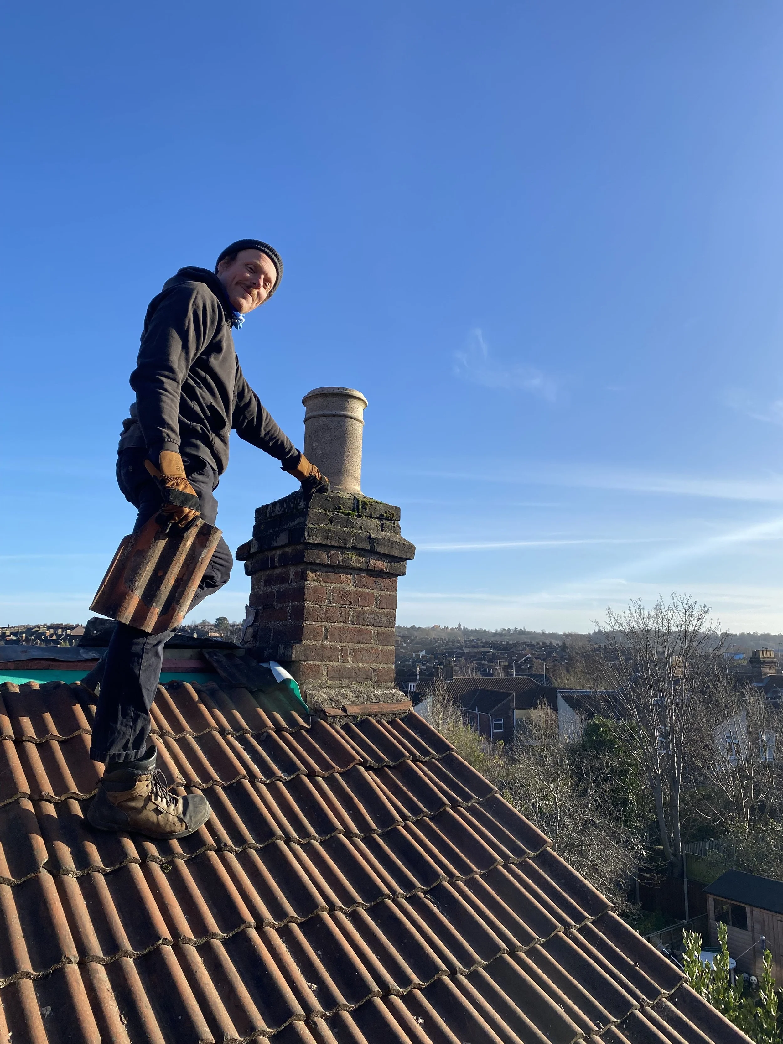 Man on a rooftop working on a chimney, holding a brick in one hand, with a clear blue sky in the background.