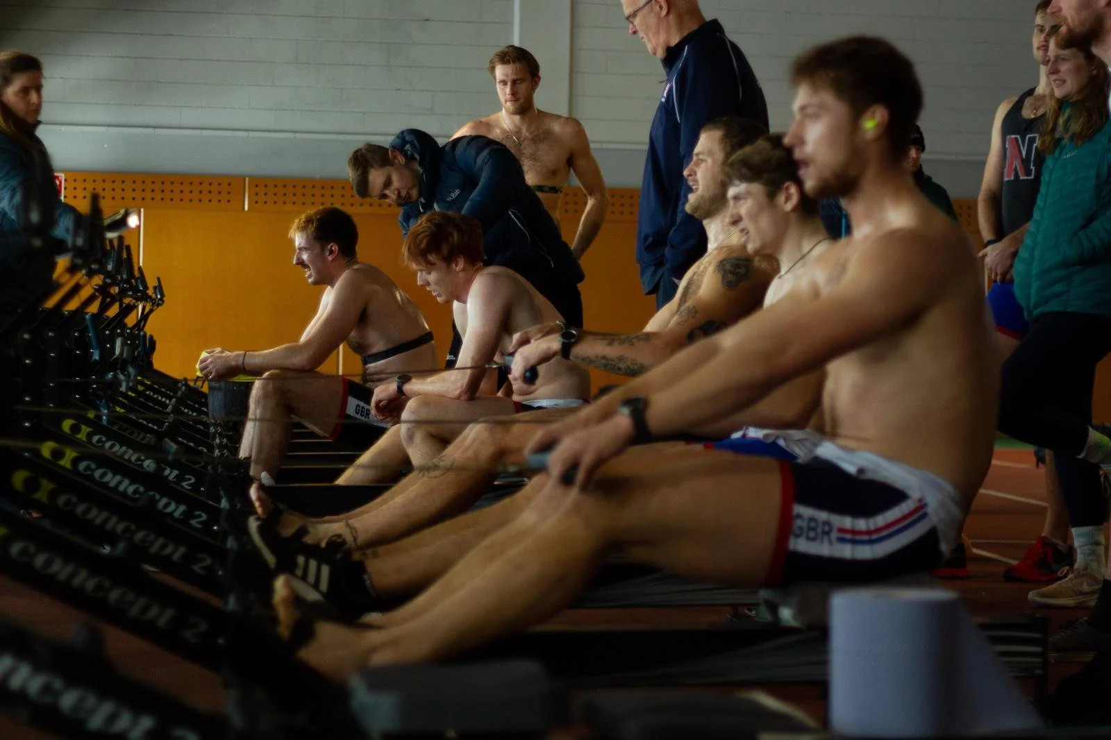 A group of male athletes at an indoor rowing competition, wearing sports shorts, using rowing machines, and being watched by other competitors and officials in the background.