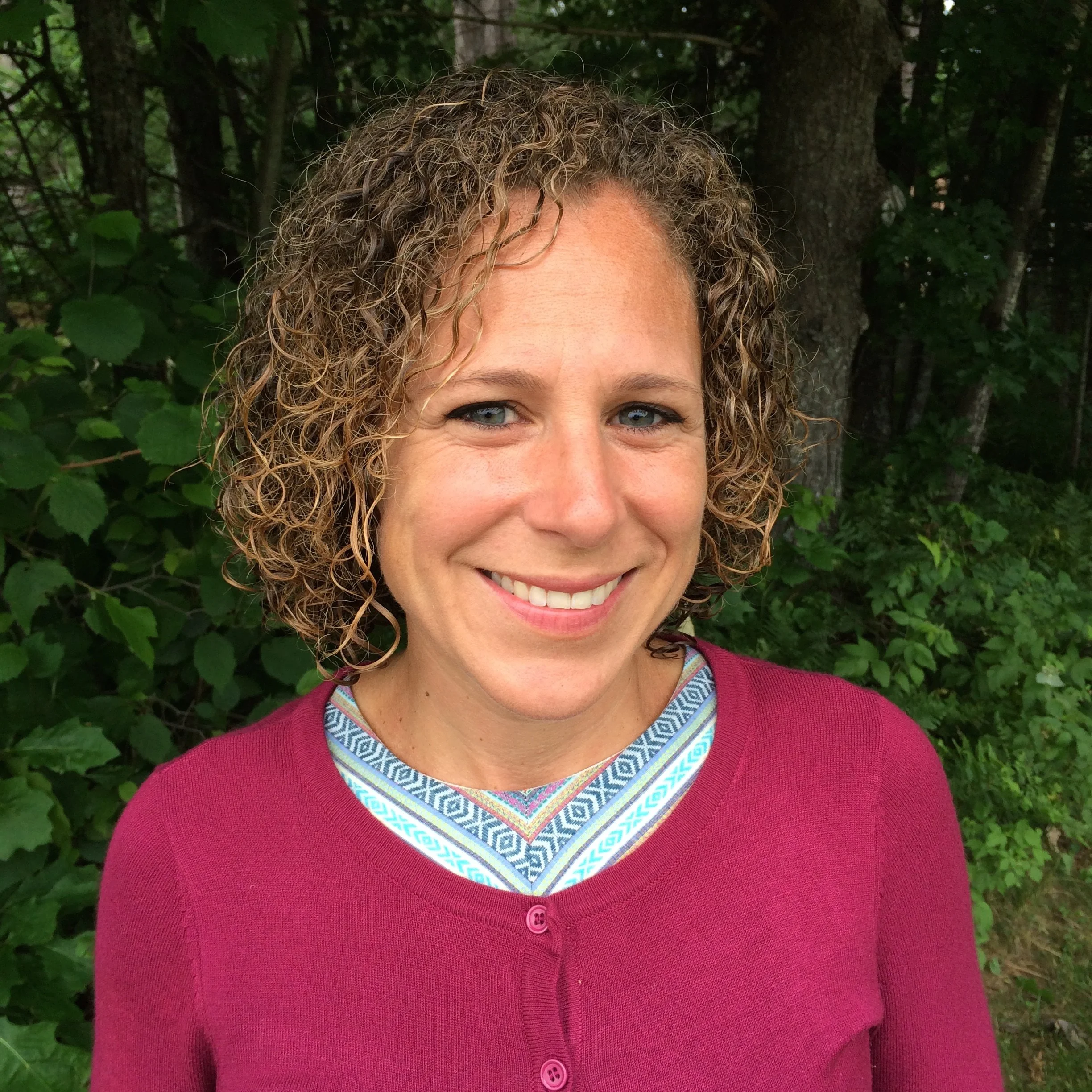 A woman with curly blonde hair and blue eyes smiling outdoors in front of green foliage, wearing a maroon cardigan over a patterned shirt.