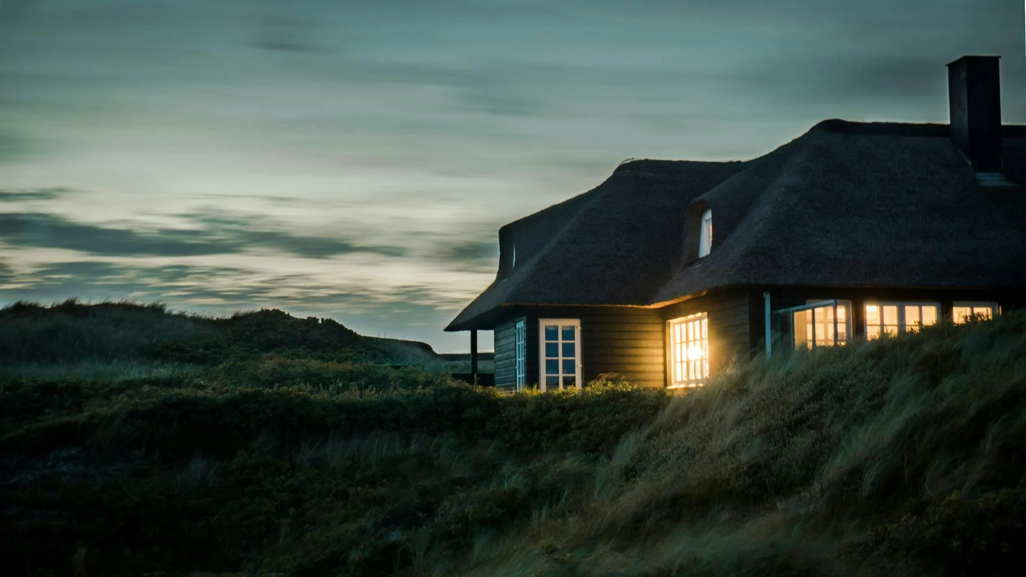 A house on a hillside at dusk with lights shining through the windows, surrounded by grassy terrain and hills under a cloudy sky.
