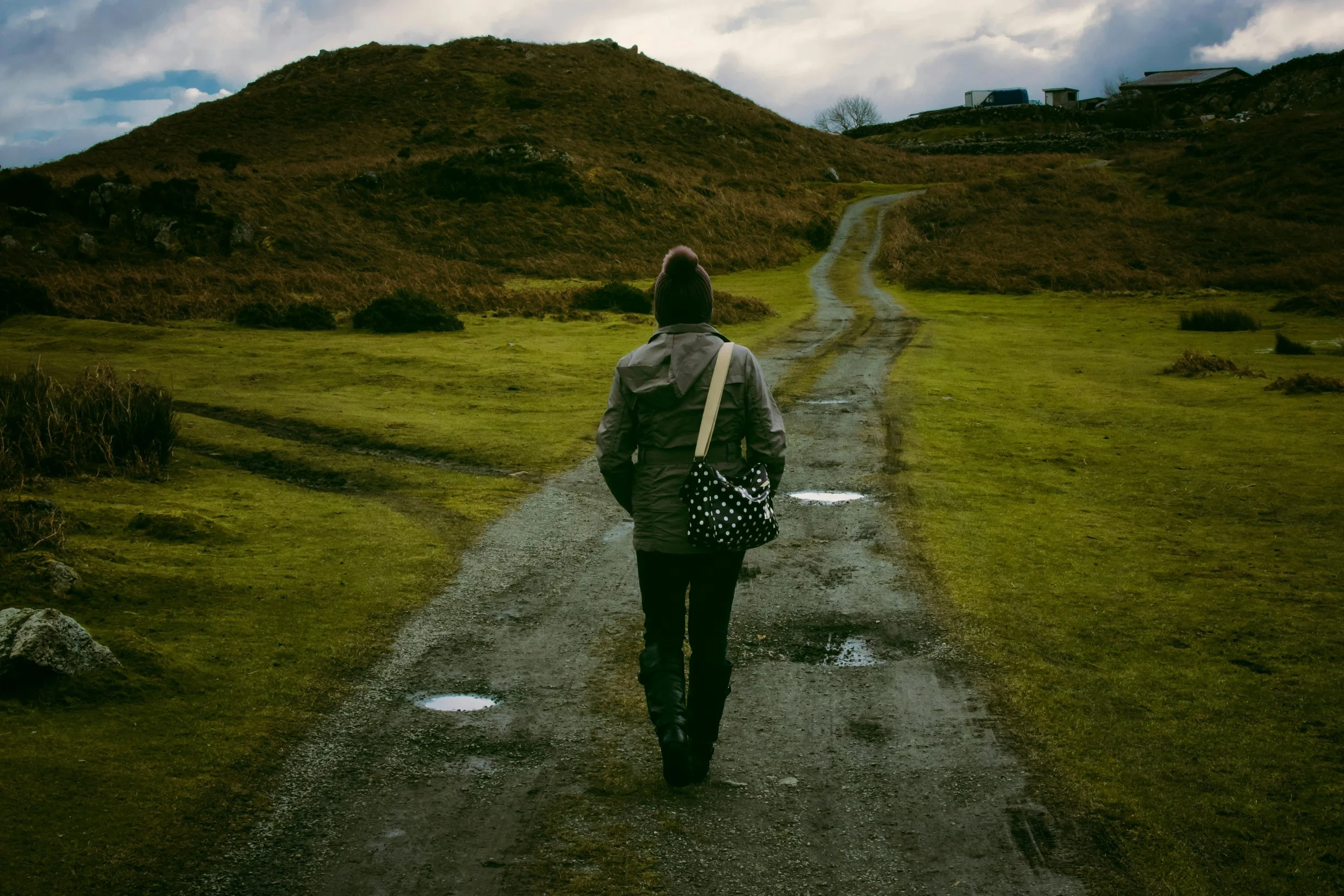 A person walking on a muddy, winding dirt road through grassy hills under a cloudy sky, carrying a polka dot bag and wearing a beanie and jacket.