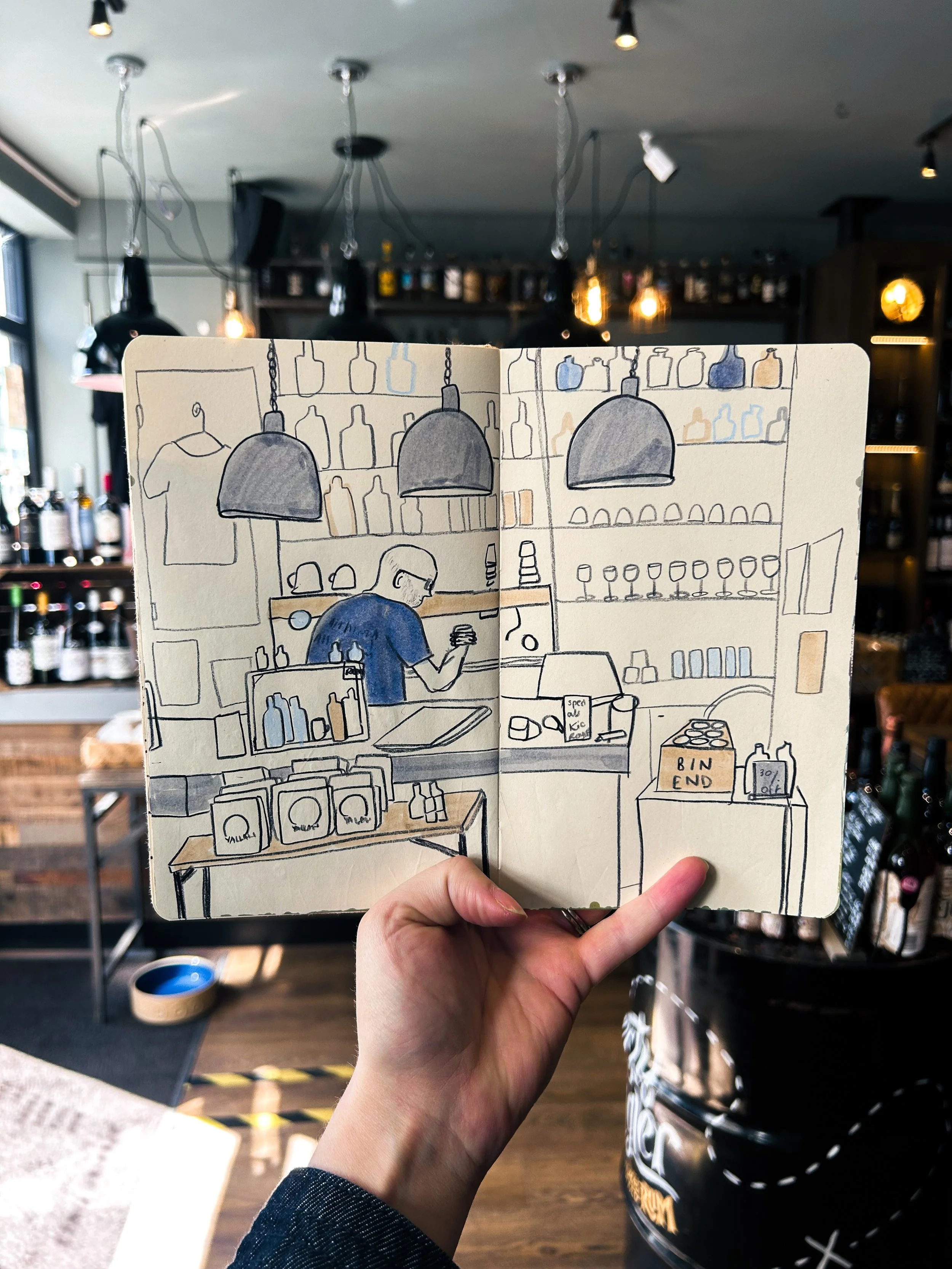 Hand holding a sketchbook with a drawing of a bar interior, showing shelves with bottles, glasses, a man behind the counter, and various bar supplies.