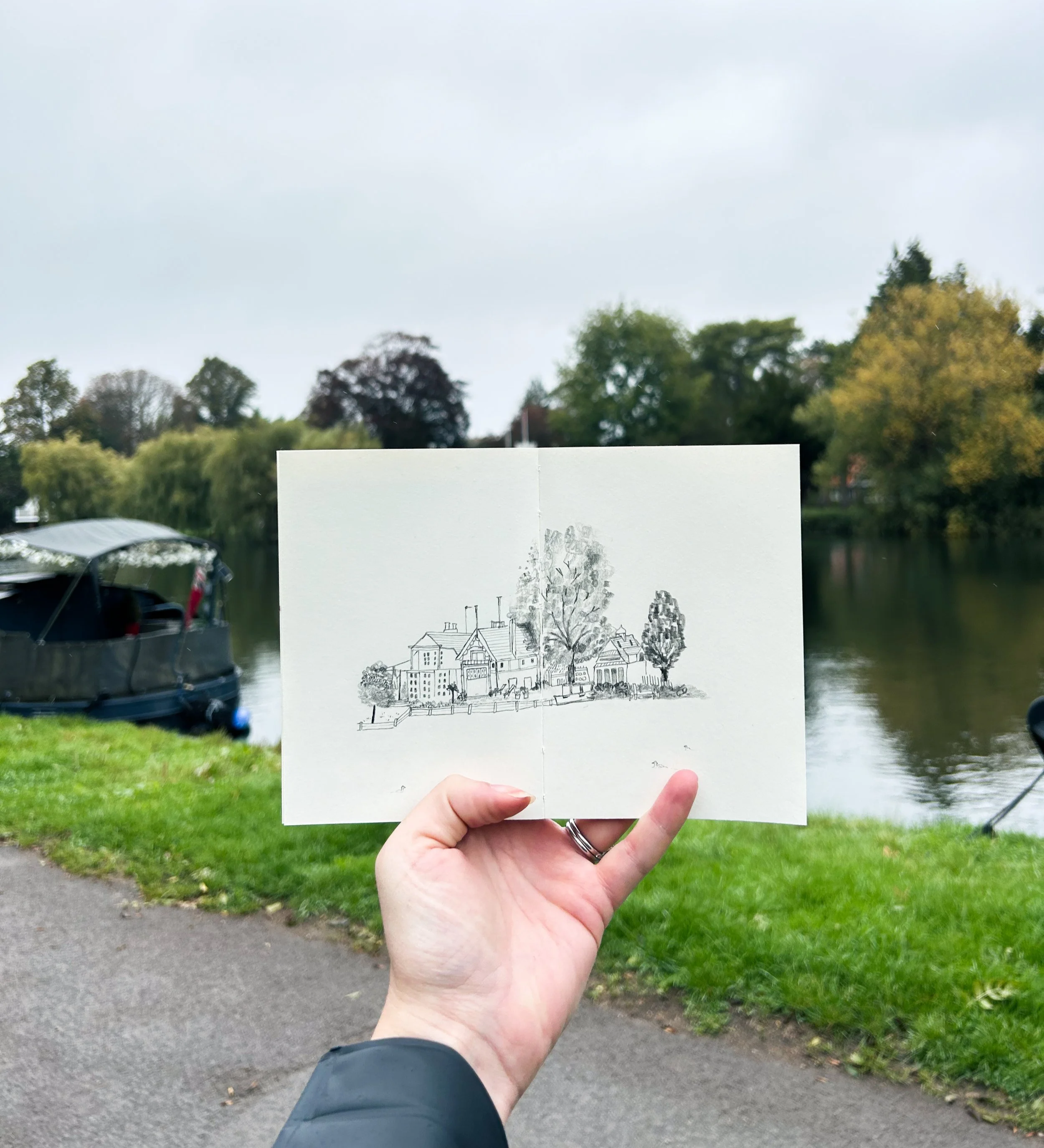 Person holding open sketchbook with drawing of houses and trees near a river, with goosenecks and boats on the water, overcast sky, trees with fall foliage in the background.
