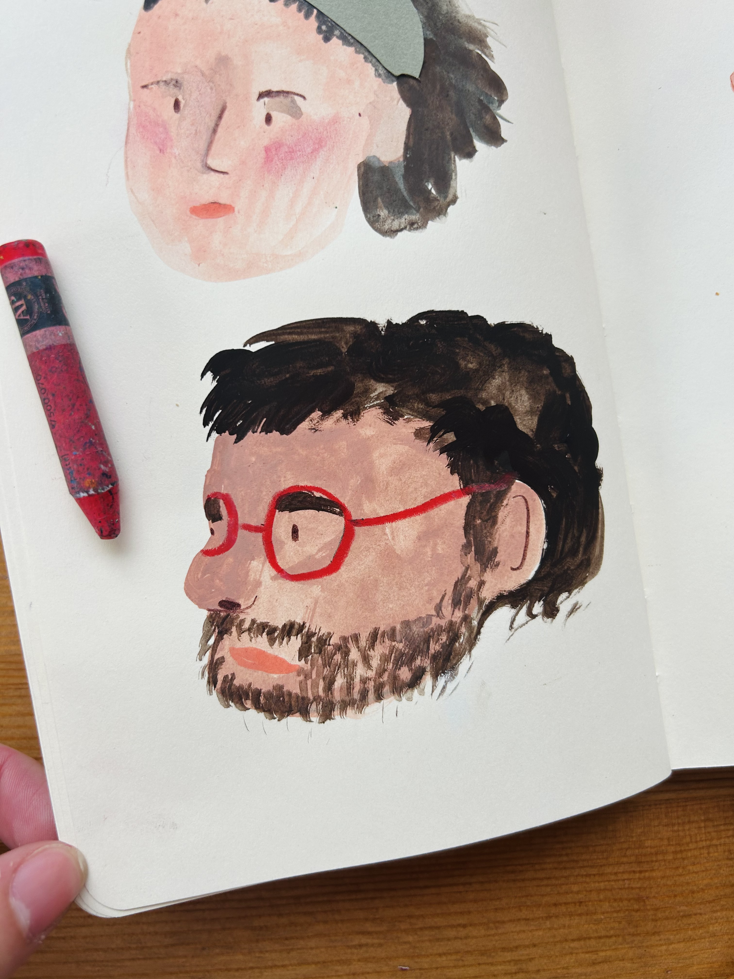 Hand-drawn watercolor portraits of a woman with short, dark hair, and a man with glasses and a beard, on a white sketchbook page with a red crayon resting beside them.
