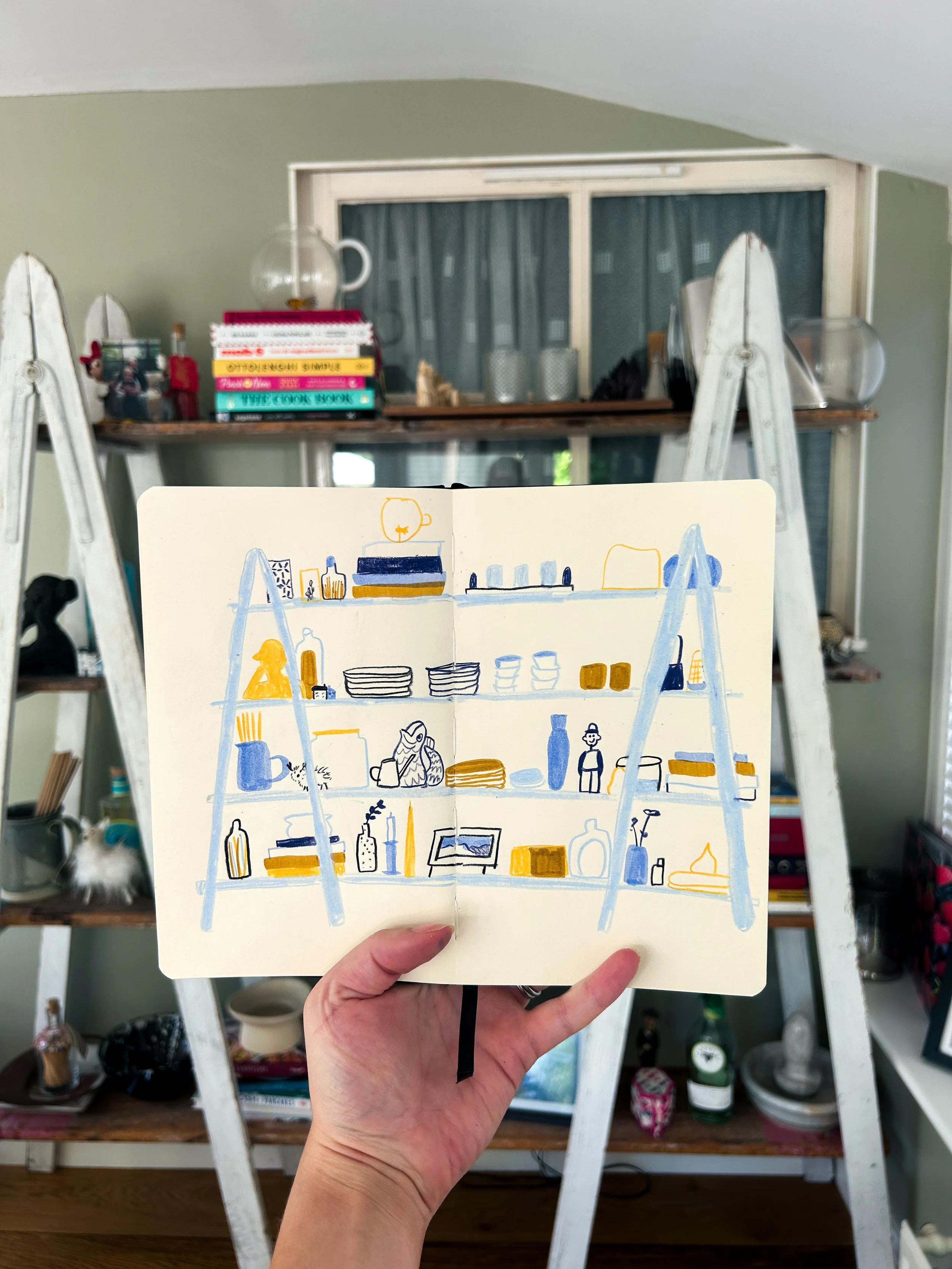 Hand holding an open sketchbook with a drawing of a shelving unit filled with books, bottles, and decorative items, with a ladder-like structure behind the sketchbook and a window with curtains above.