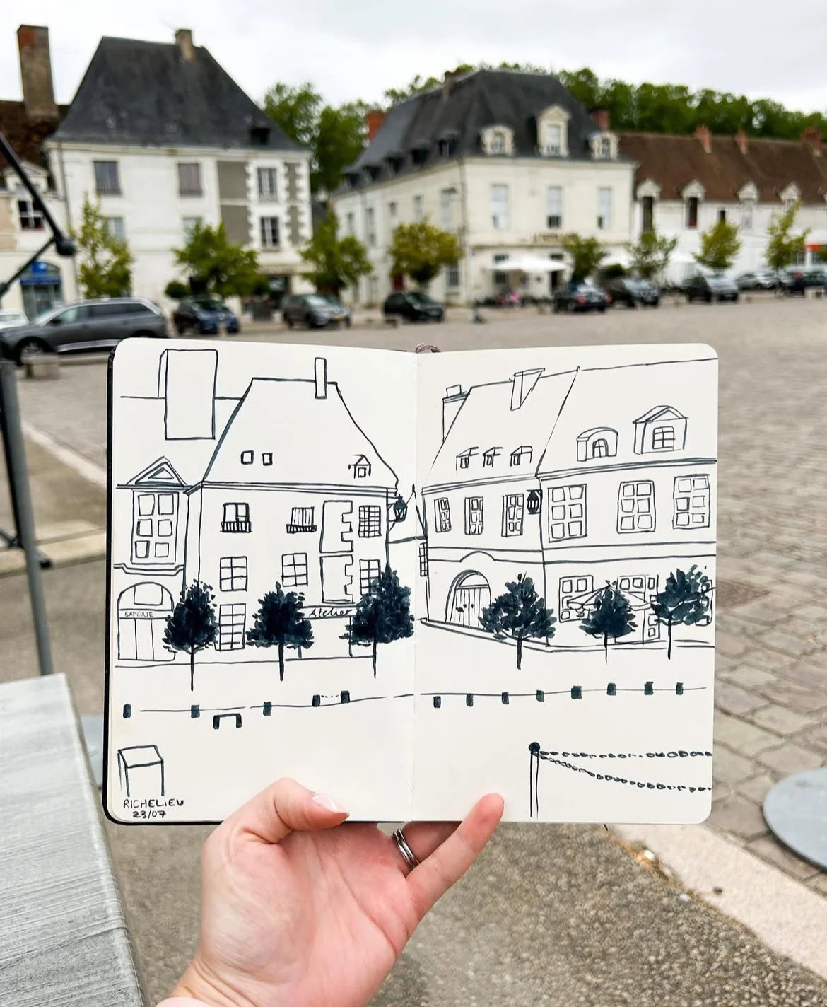 Hand holding a sketchbook with a drawing of buildings, trees, and a chain in front of a row of existing buildings and parked cars in a European town square.