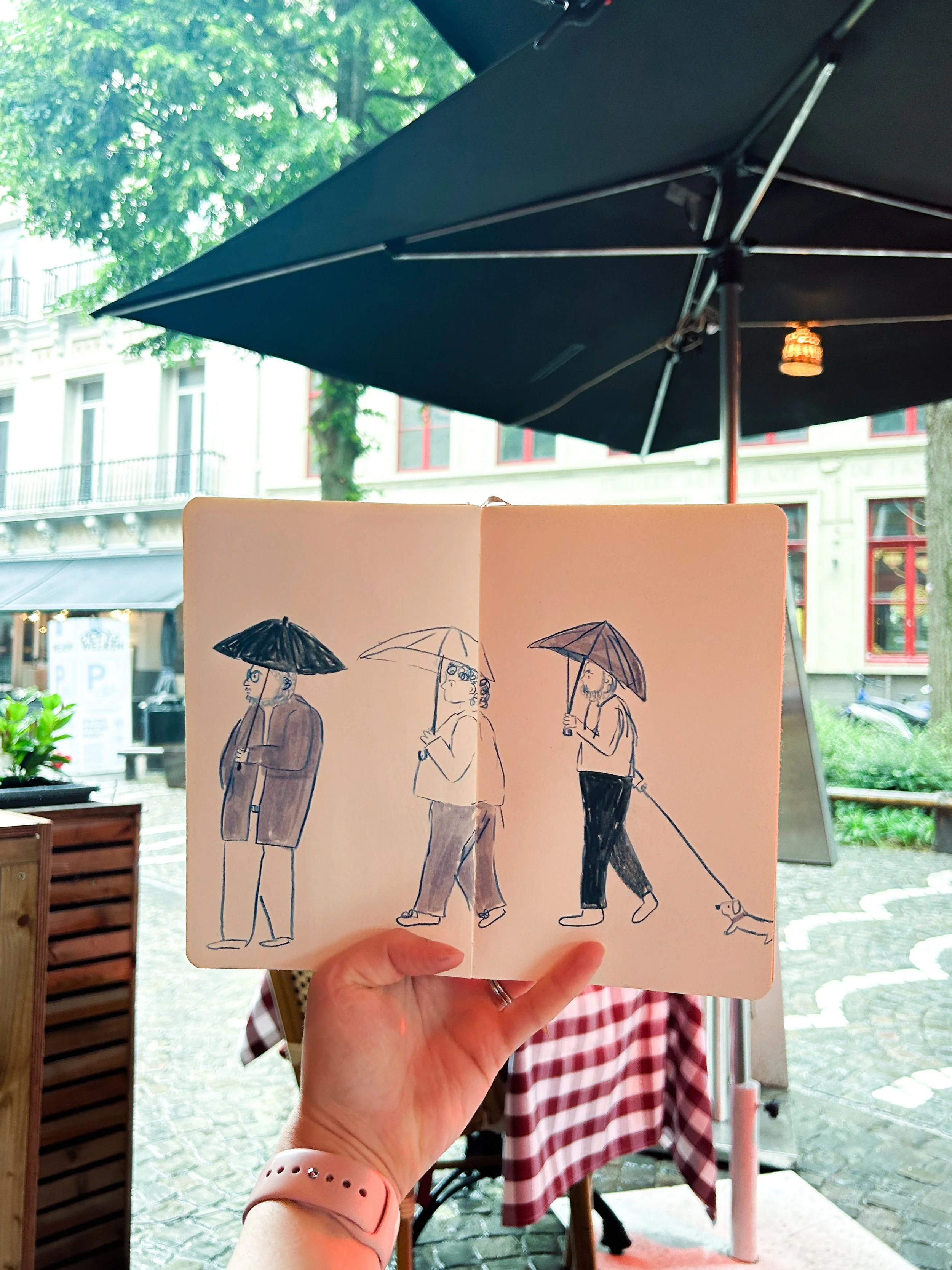 Hand holding an open sketchbook with a drawing of three people walking with umbrellas, while outdoors under a patio umbrella.