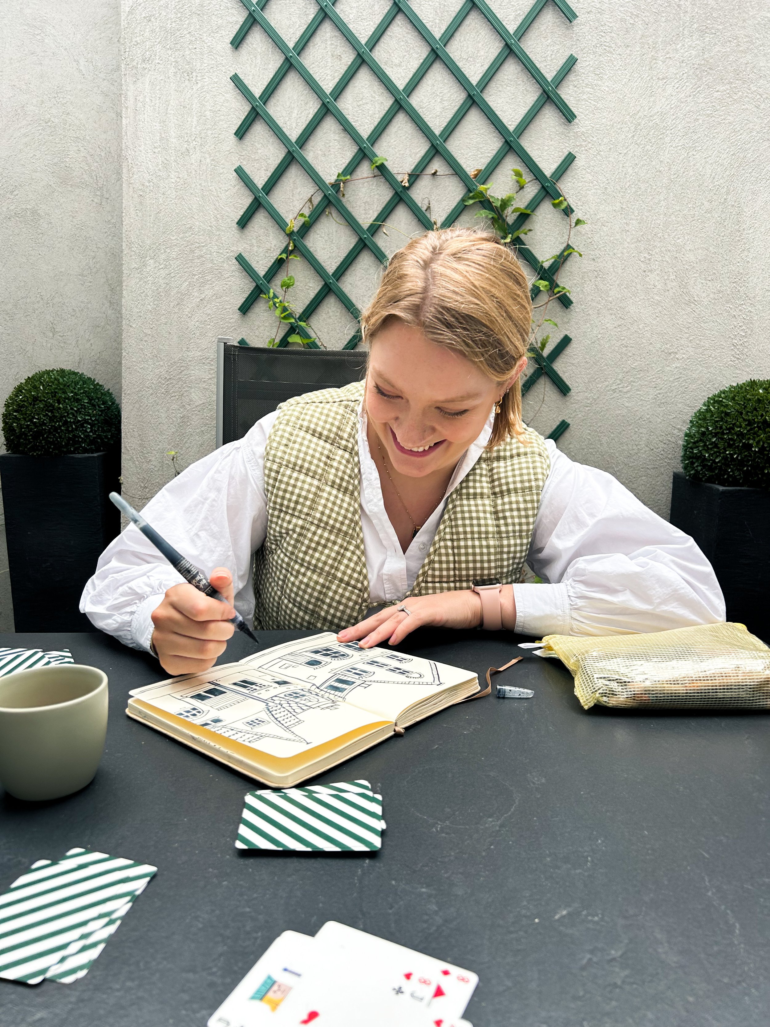 A woman smiling while drawing house sketches in a notebook at an outdoor table with a cup, playing cards, and a packet of tea or snacks, with a green trellis and potted plants in the background.