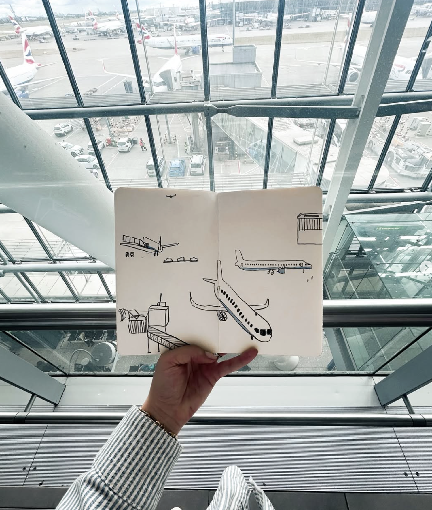 A person holding a sketchbook with drawings of airplanes at an airport terminal with aircraft parked outside the windows.