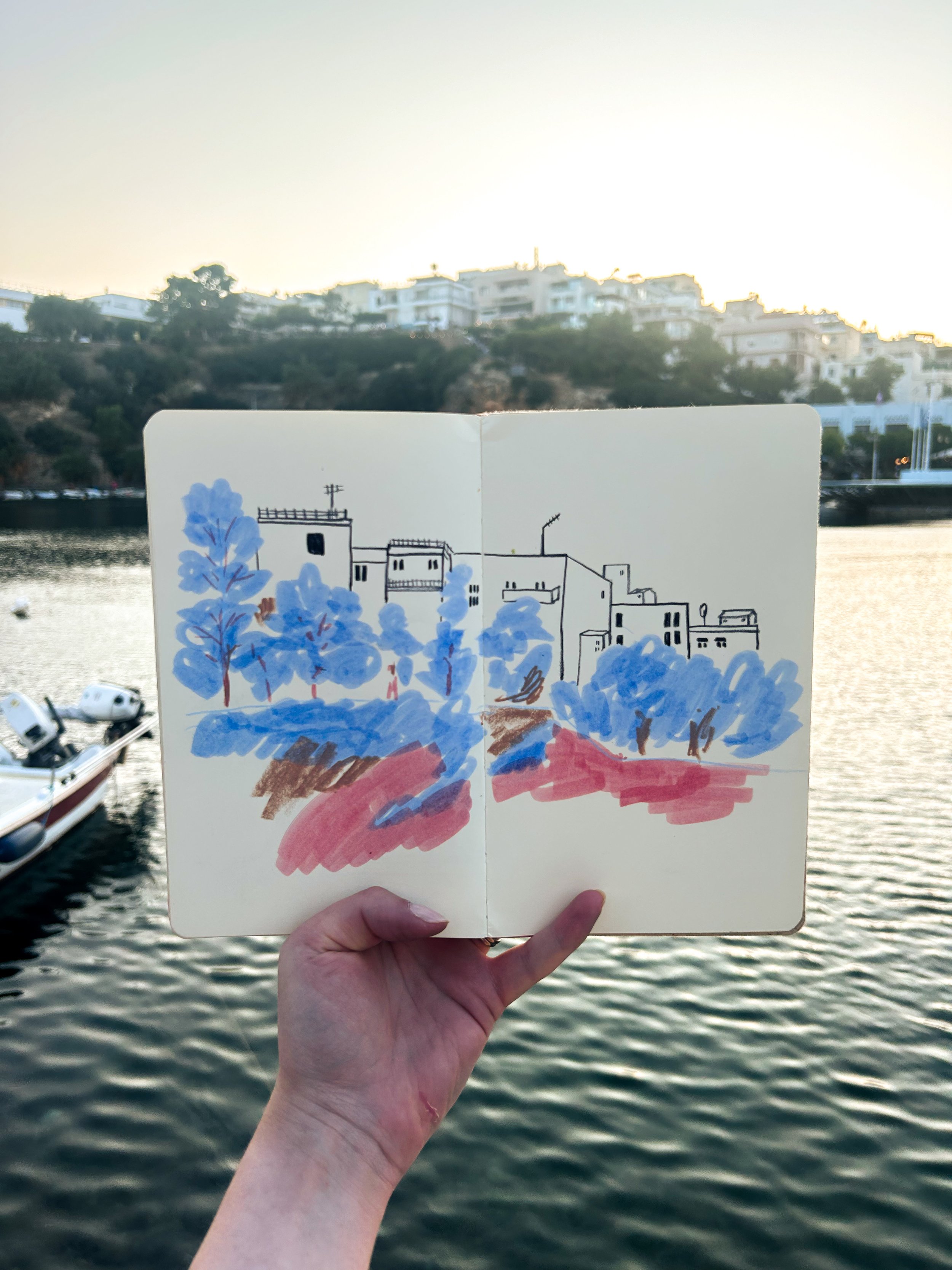 A hand holding an open sketchbook with a colorful drawing of trees and buildings, held against a backdrop of a body of water, boats, and a hillside with white buildings at sunset.