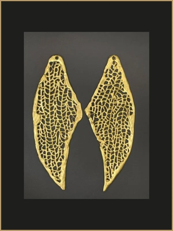 Deux pendentifs en forme de feuille en métal doré avec un motif de grille complexe, posés sur un fond noir.