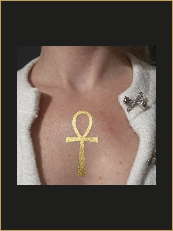 Tatuage d'une ankh dorée sur la peau du décolleté d'une femme, portant un vêtement blanc avec un bouton en forme de nœud en métal.