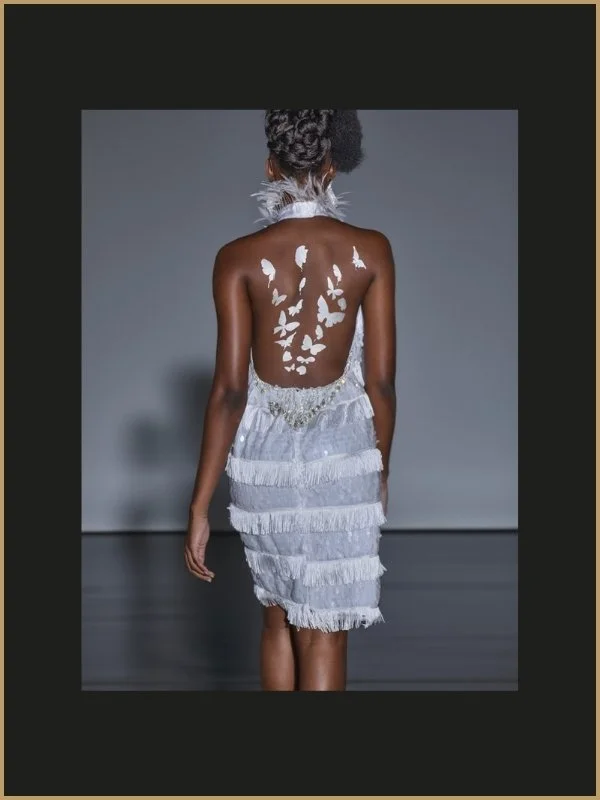 Une femme marchant sur un podium, vue de dos, portant une robe blanche avec des franges et des détails brillants, avec des papillons blancs apposés sur son dos.