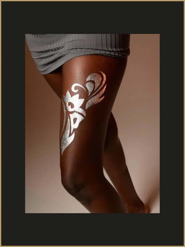 Une cuisse avec un tatouage tribal argenté sur la peau foncée, une personne portant un vêtement gris
