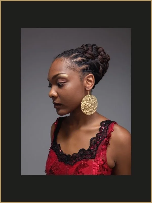 Une femme avec des cheveux coiffés en dreadlocks et portant une robe rouge avec des détails en dentelle noire, portant une grande boucle d'oreille en or circulaire.