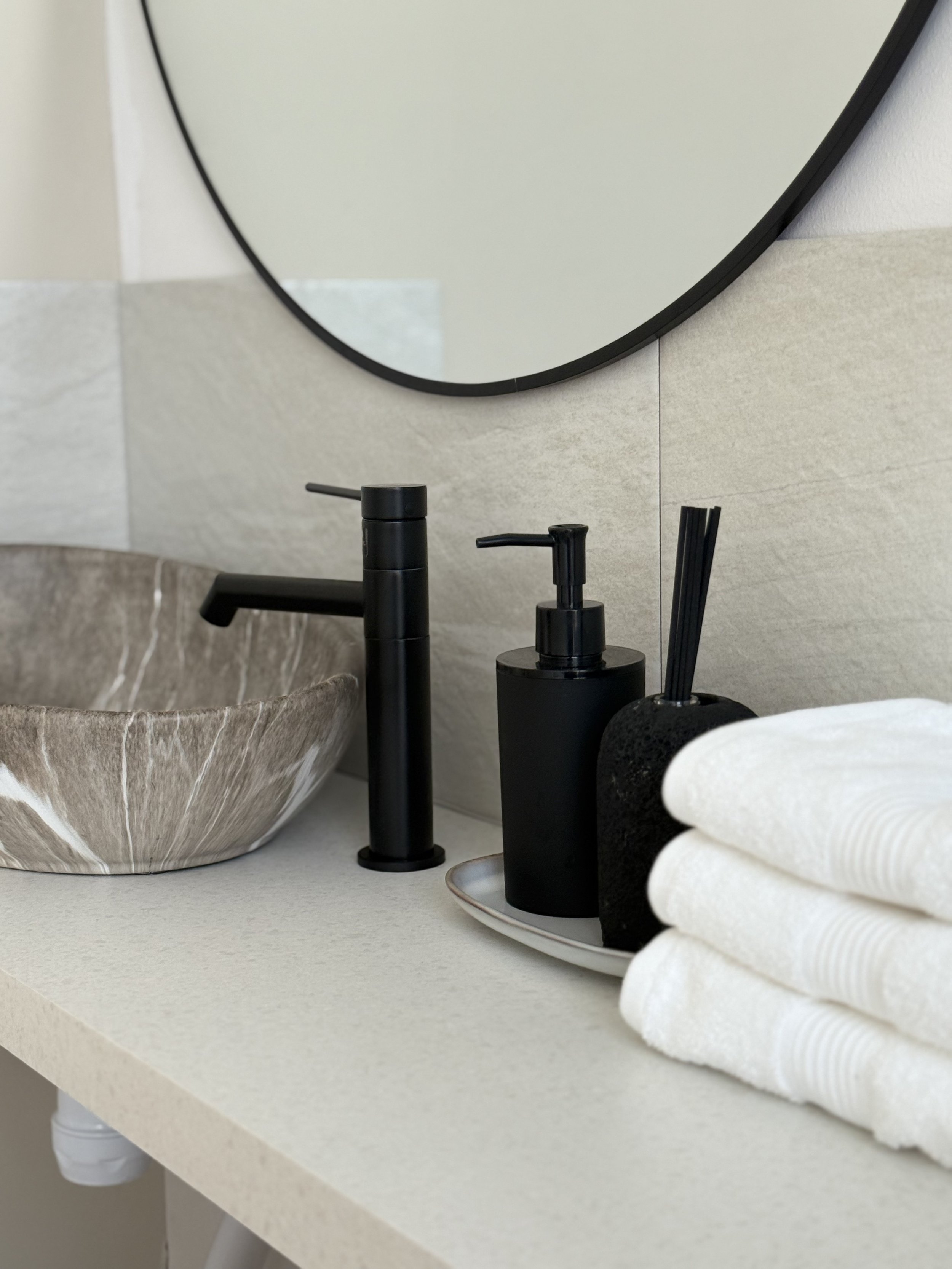 Ett badrumsspel med en naturstensfärgad vask, svart handfatkran, två svarta tvålpumpar, en svart borste, en spegel och vita handdukar.