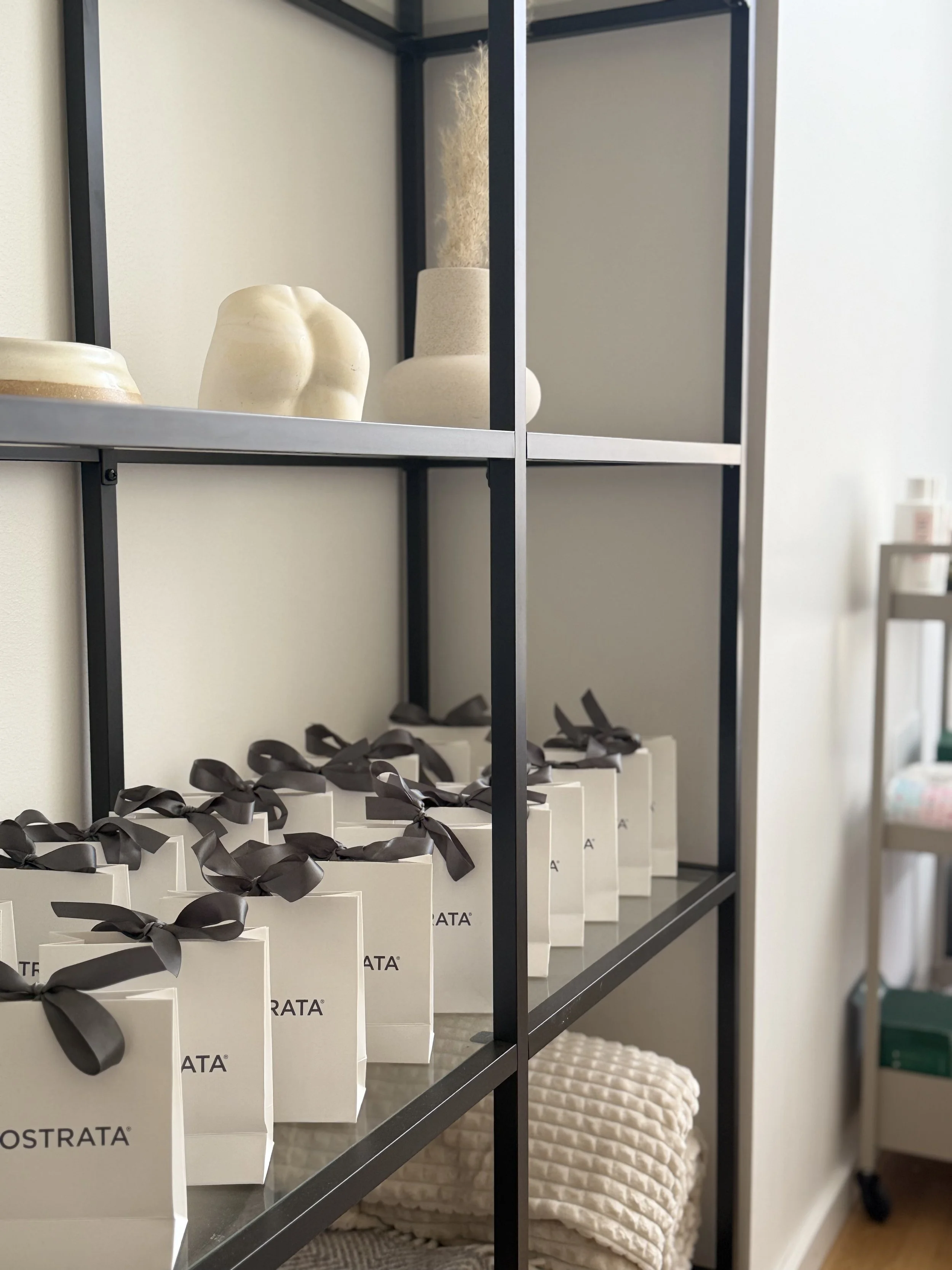 Vita presentförpackningar med svarta satinband placerade på en svart metallhylla, under en vit stickad skulltavla med runda mönster. Bakgrund med ytterligare hyllor och ett vitt vägg.