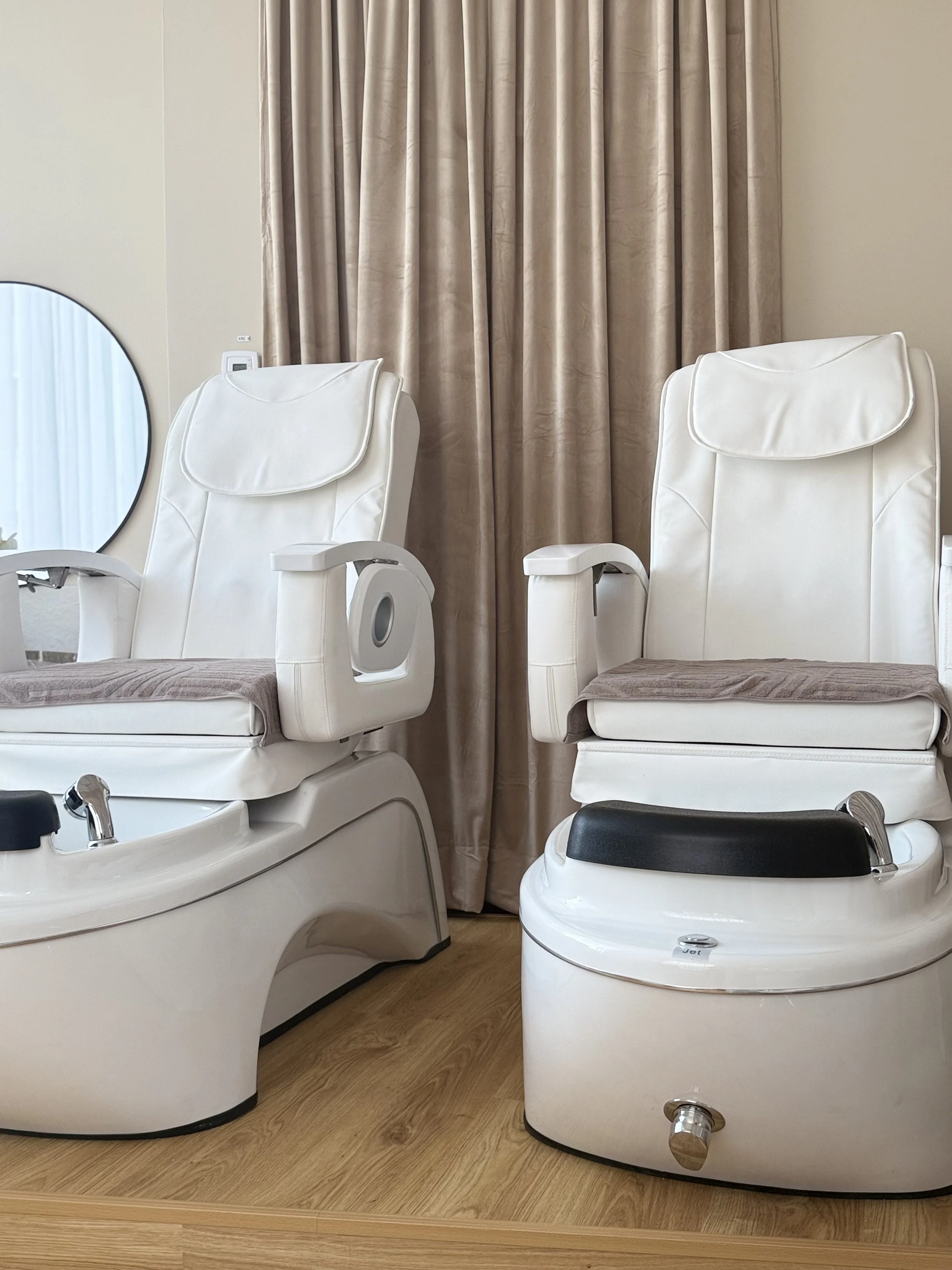 Två vita pedikyr- eller fotbad i ett rum med beige väggar, beige gardiner och trägolv.