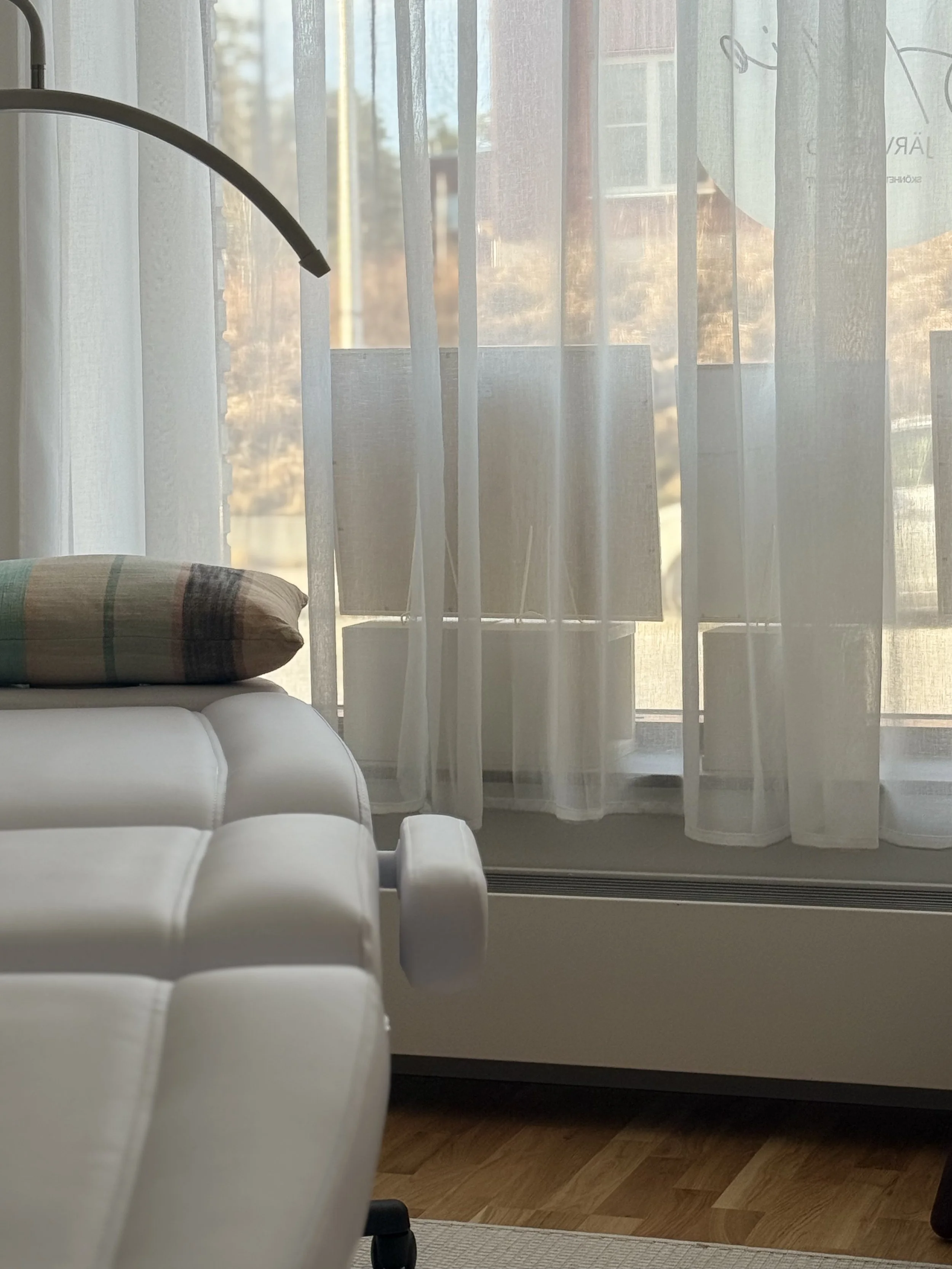 En vit soffa med en kudde som har en randig mönster i beige, grönt, svart och rött, placerad framför ett fönster med genomskinliga vita gardiner.