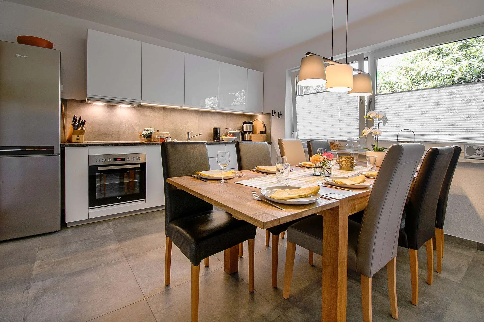 Moderne Küche mit Essbereich, gedecktem Tisch, weißen und schwarzen Stühlen, großen Fenstern mit Rollos, moderner Beleuchtung