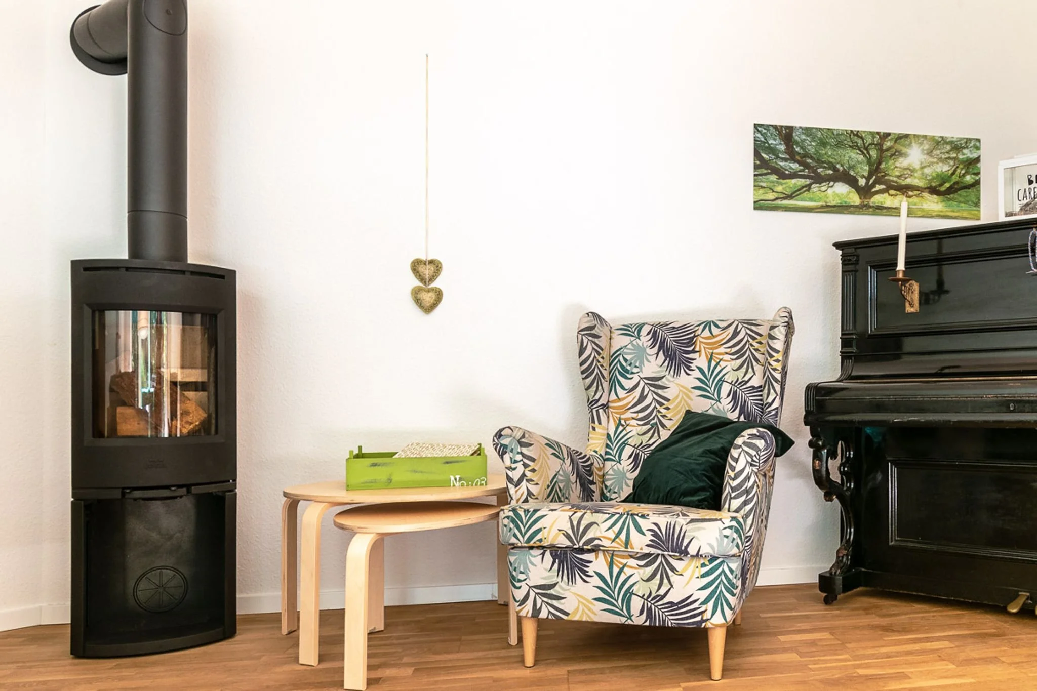 Gemütliches Wohnzimmer mit schwarzen Kamin, einem armlehnensessel mit Blattprint, einem kleinen Beistelltisch mit grüner Kiste, einer schwarzen alten Schrank, Wanddeko in Herzform und einem Bilderrahmen mit Baumaufnahme.