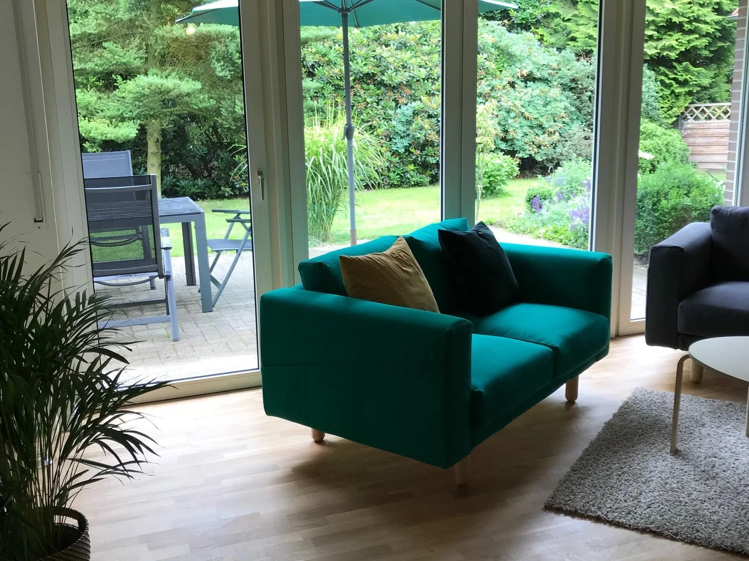 Innenansicht eines Wohnzimmers mit grüner Couch, Kissen, einem kleinen Tisch, einem Teppich und Blick auf einen Garten mit Terrasse, Stuhl und Sonnenschirm.