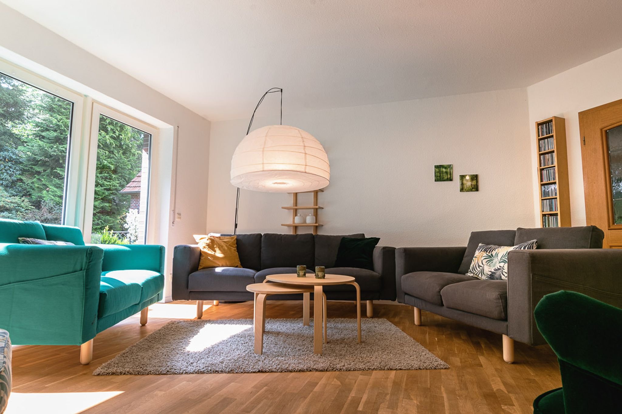 Wohnzimmer mit grauen und grünen Sofas, Holztisch, Teppich, großer Fenster, hängende Papierlampe, Wanddekoration mit kleinen Bildern, CD-Regal, Holztür.