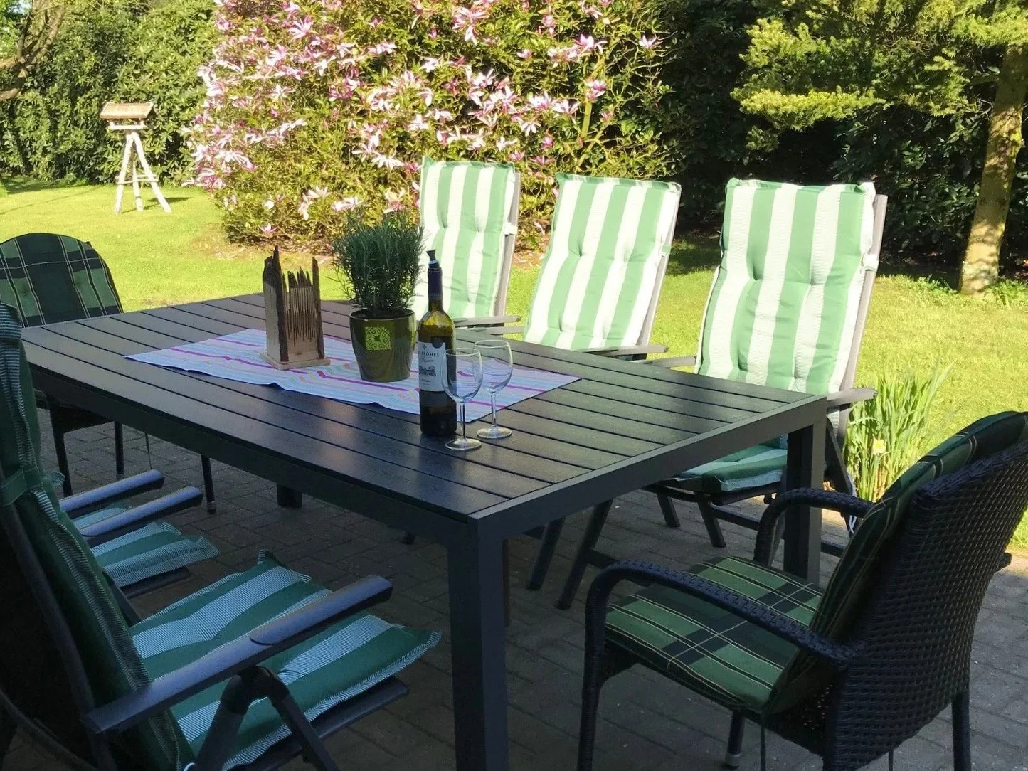 Gartenmittagstisch mit Wein, Gläsern, Blumen und Stühlen im Garten.