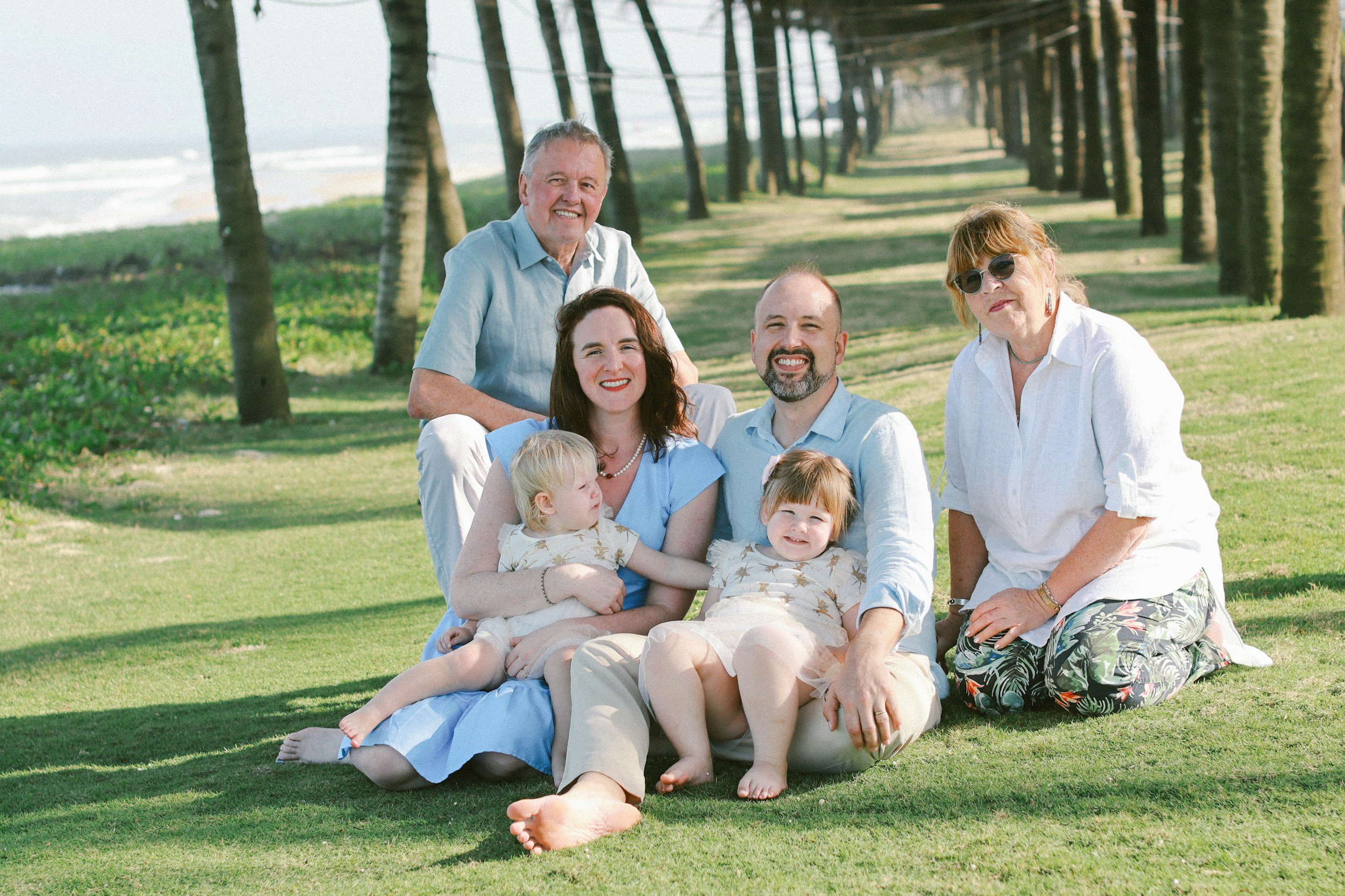 A multigenerational family of six people sitting on the grass under a row of palm trees on a sunny day at the beach.