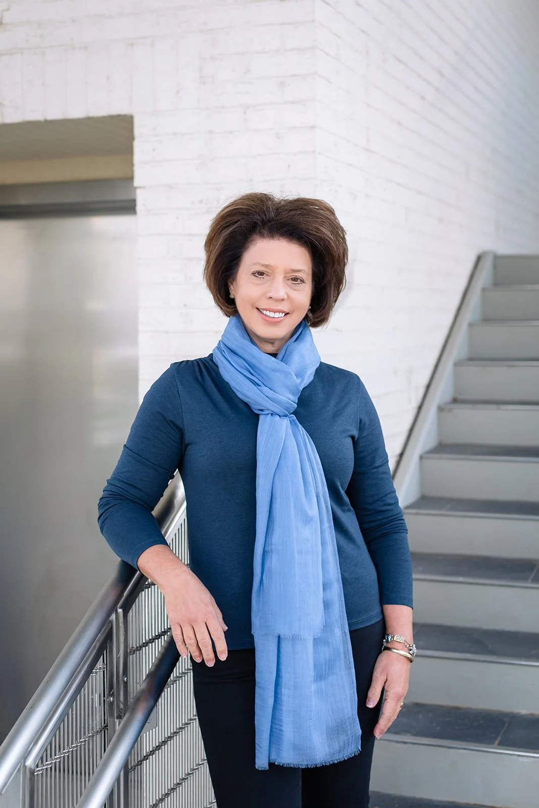 A smiling woman in a dark blue top with a light blue scarf, standing in front of a white brick wall with stairs in the background.