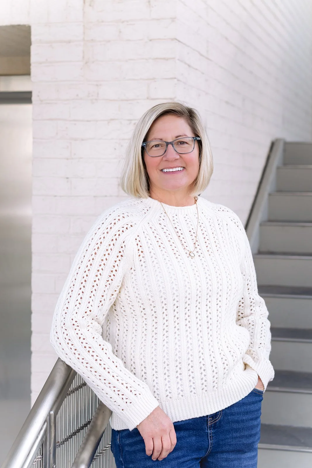 A woman with blonde hair, glasses, and a white knit sweater standing against a white brick wall and a metal staircase, smiling at the camera.