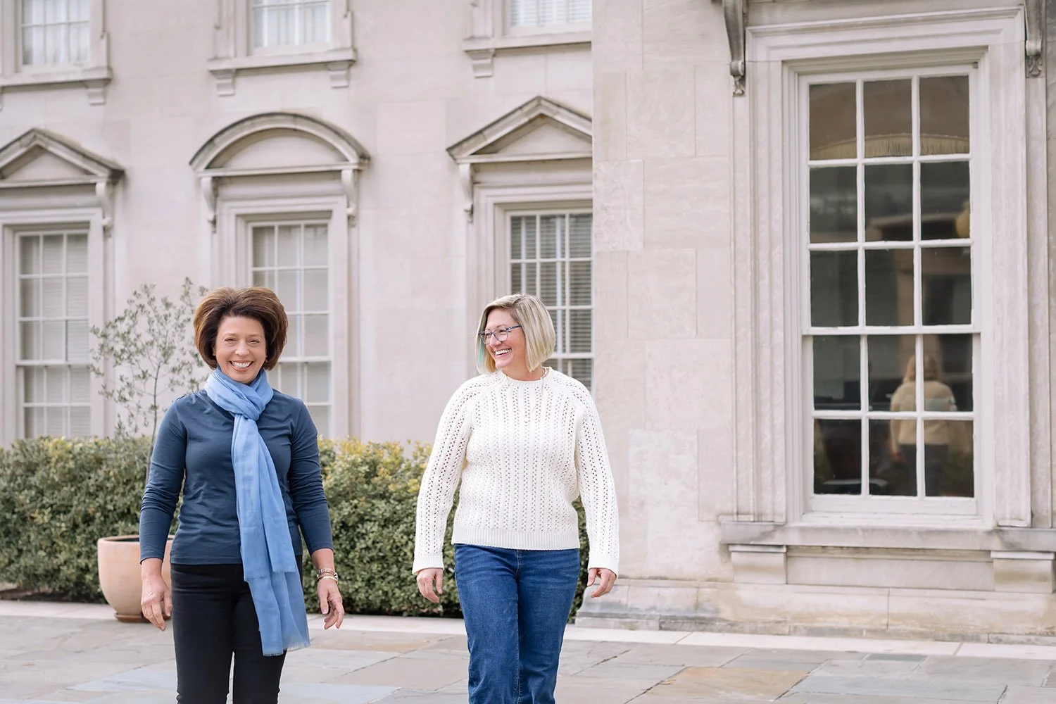 Two women walking and smiling outside a building with large windows and stone facade.