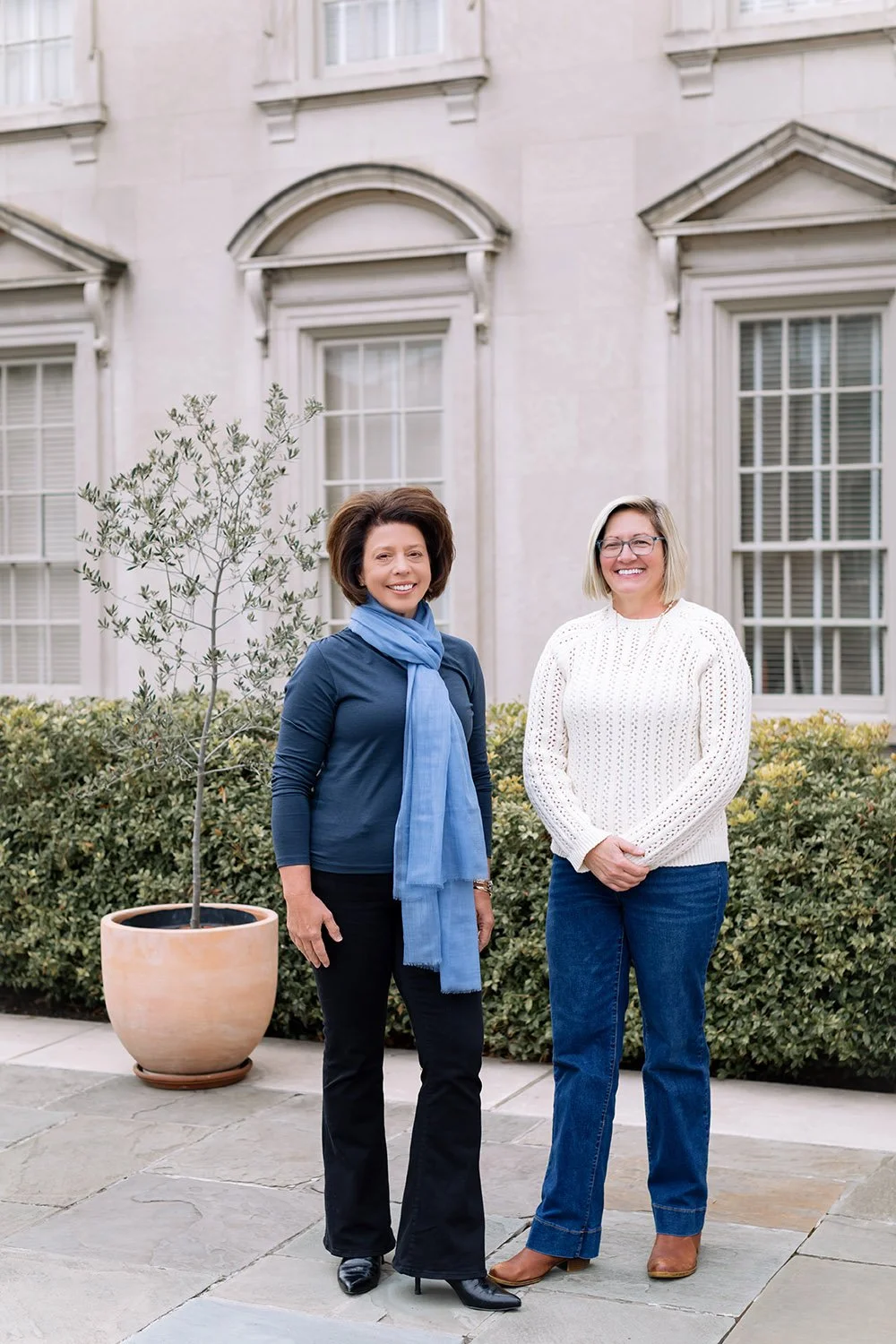 Two women standing outdoors in front of a building with large windows and plants, smiling at the camera.