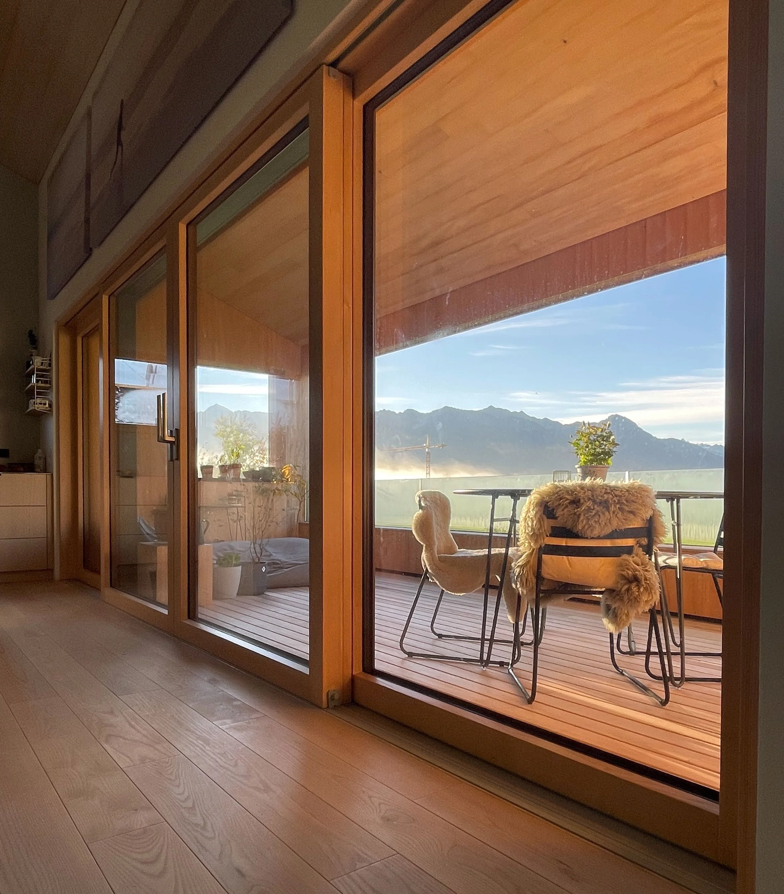 Innenraum mit großen Glas-Schiebetüren, die auf eine Holzterrasse mit einem Tisch und Stühlen vor einer Bergkulisse hinausführen.