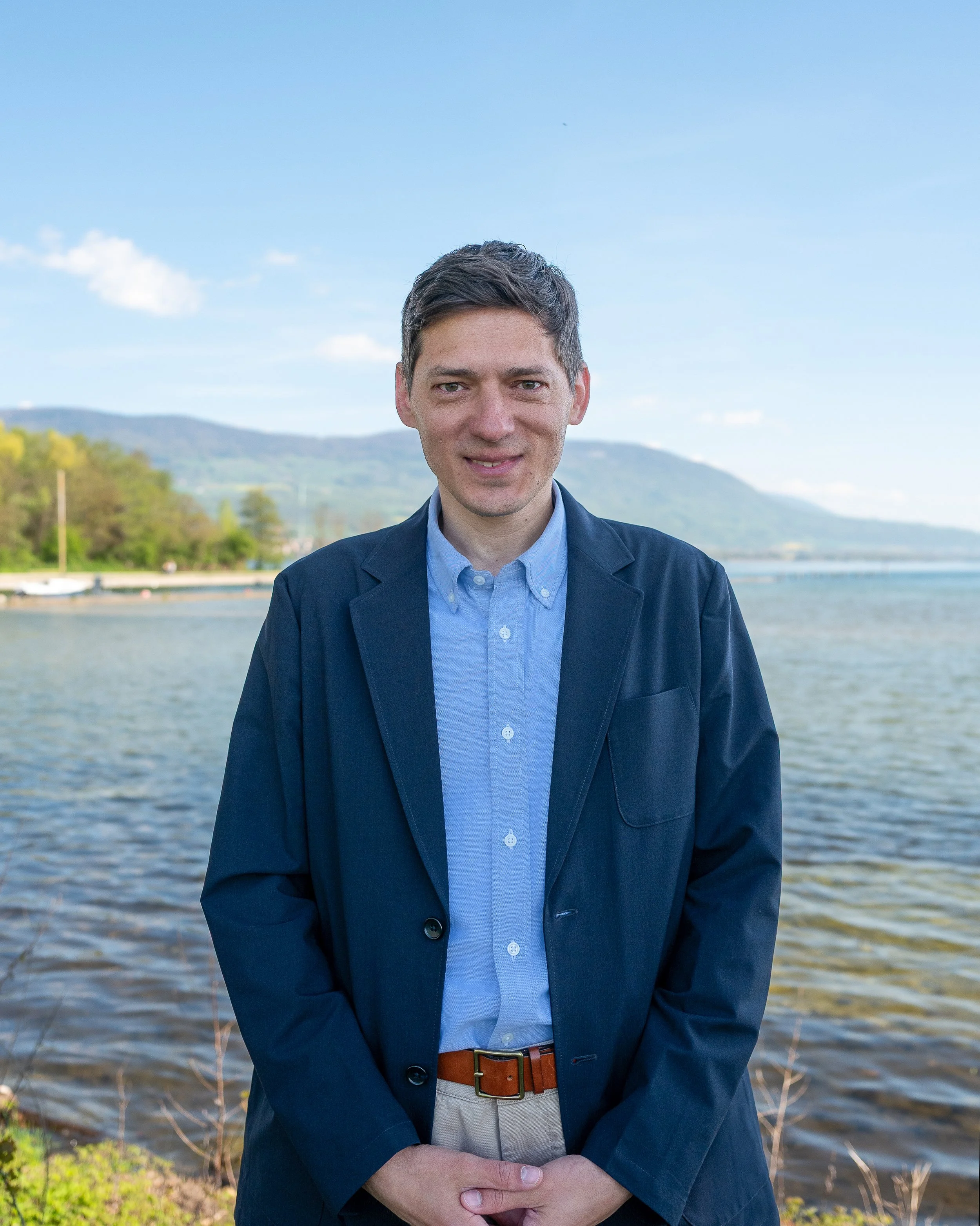 Un homme en veste sombre, chemise bleue et ceinture marron se tient devant un lac avec des montagnes et un ciel bleu en arrière-plan.
