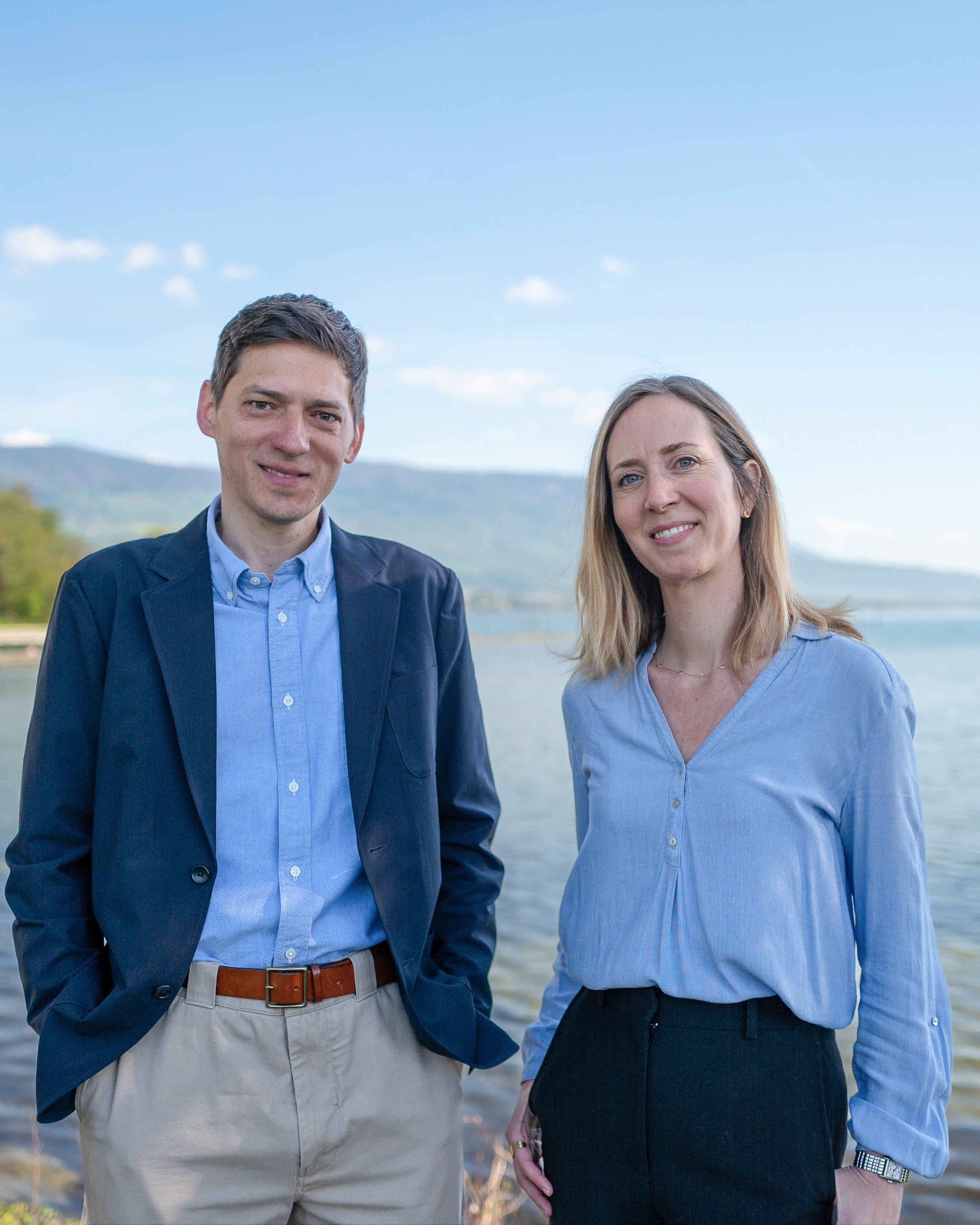 Un homme et une femme posent côte à côte devant un lac ou une rivière, avec des montagnes à l'arrière-plan, par une journée ensoleillée.