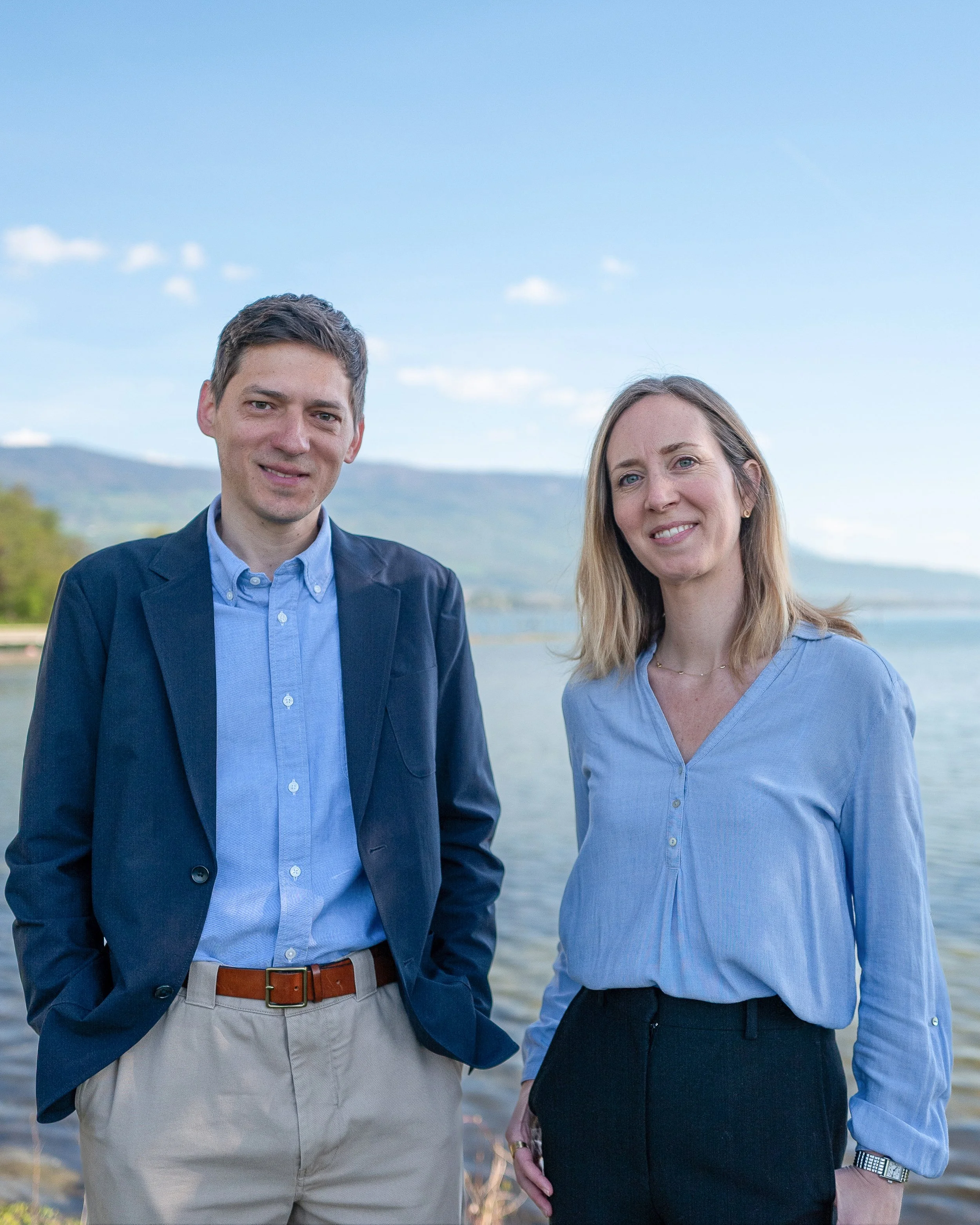 Un homme et une femme se tiennent côte à côte près d'un lac avec des montagnes en arrière-plan, tous deux souriants.