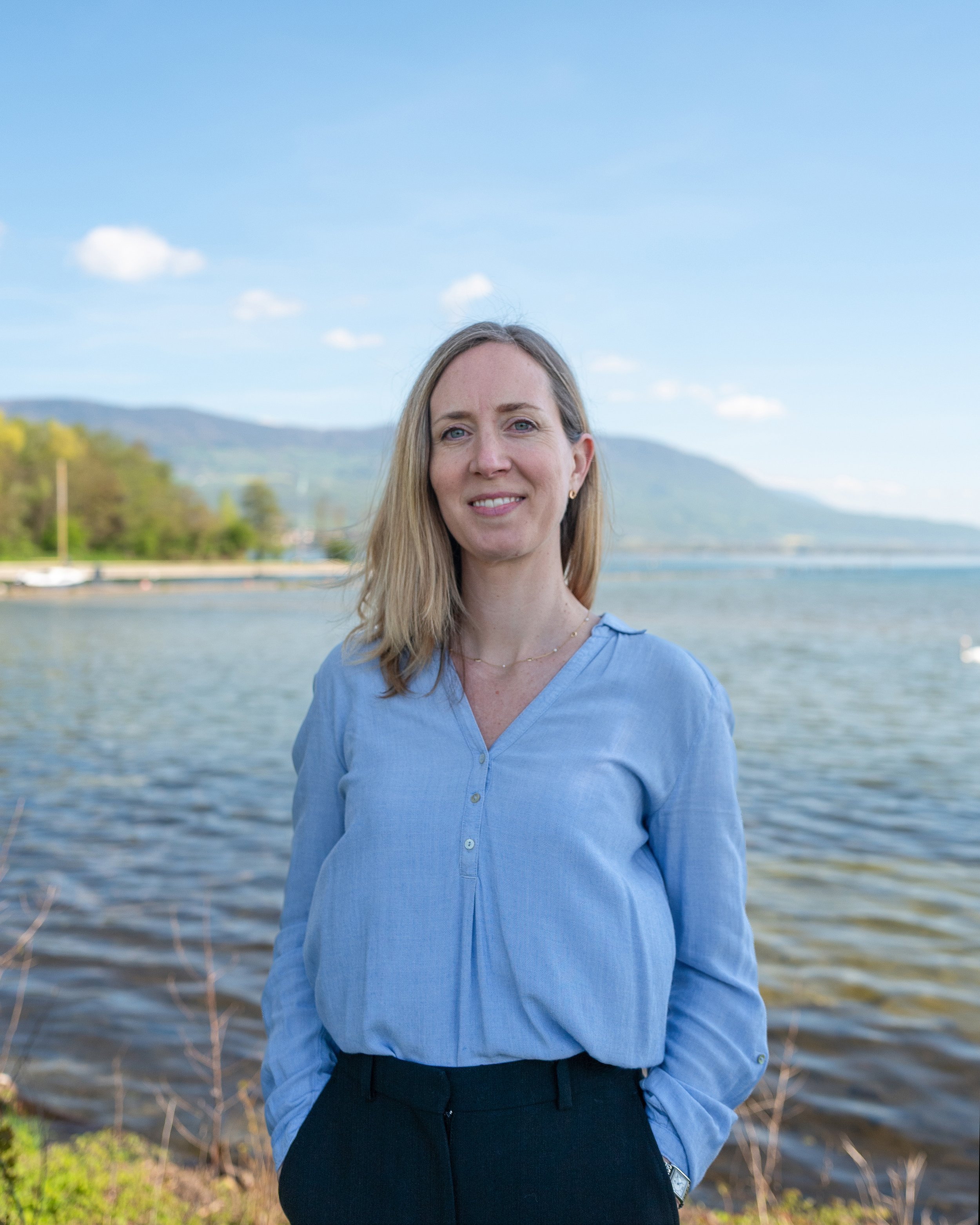 Jeune femme souriante portant une chemise bleue, debout près d'un lac avec des montagnes en arrière-plan, en pleine journée ensoleillée.