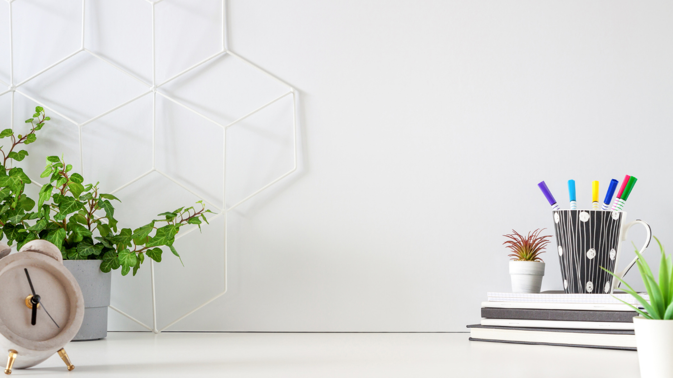 Minimalist white desk with small potted plants, a stack of notebooks, and a black mug filled with colored pens, with a decorative wall piece in the background.