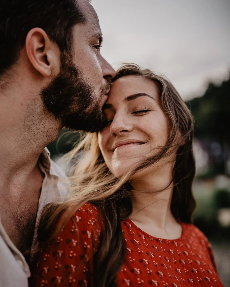 Ein Mann küsst eine lachende Frau auf die Stirn, beide sind im Freien, der Himmel ist bewölkt.