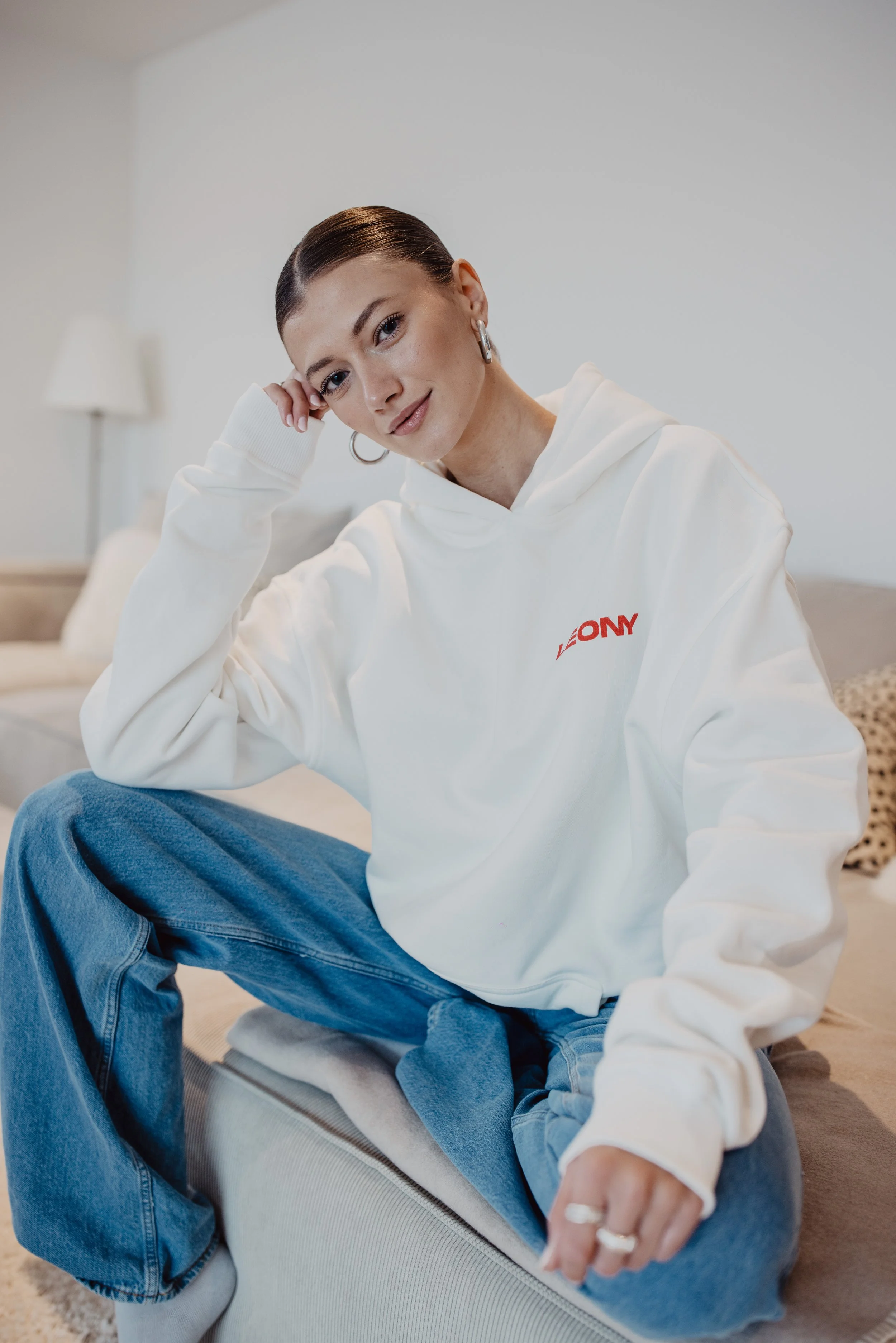 Eine junge Frau mit braunen Haaren, die einen weißen Kapuzenpullover trägt mit rotem Schriftzug, sitzt auf einem Hocker in einem hellen Raum, lächelt und schaut in die Kamera.