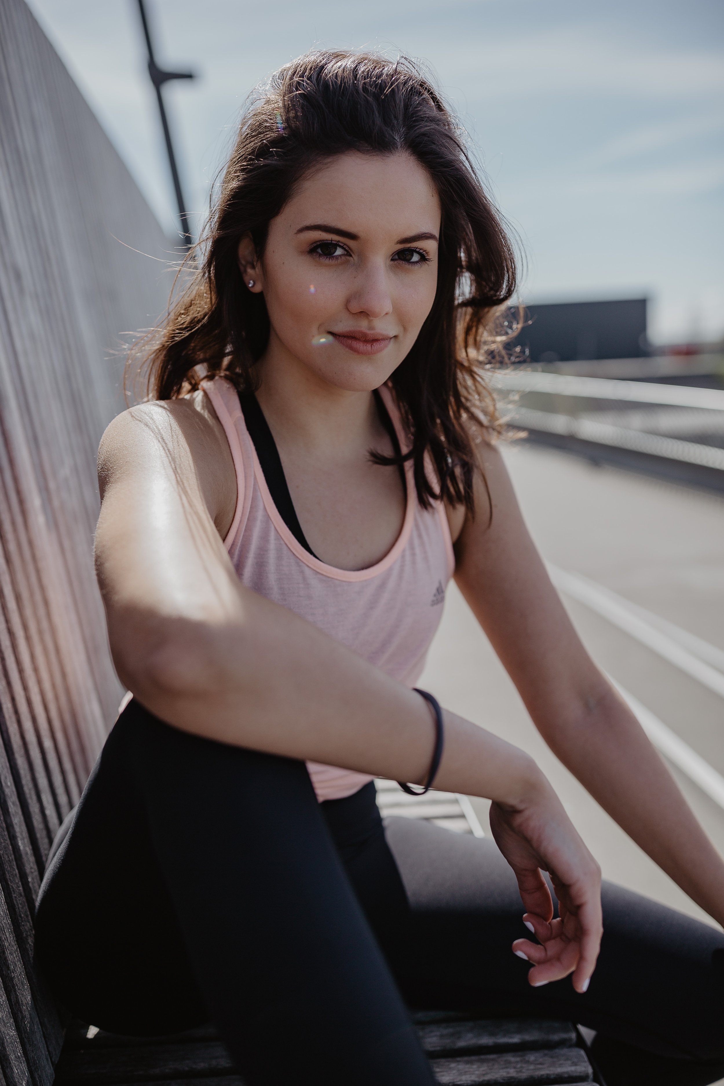 Eine junge Frau sitzt auf einer Bank im Freien, trägt ein pinkes Sporttop und schwarze Leggings, im Hintergrund ist ein blauer Himmel mit wenigen Wolken zu sehen.