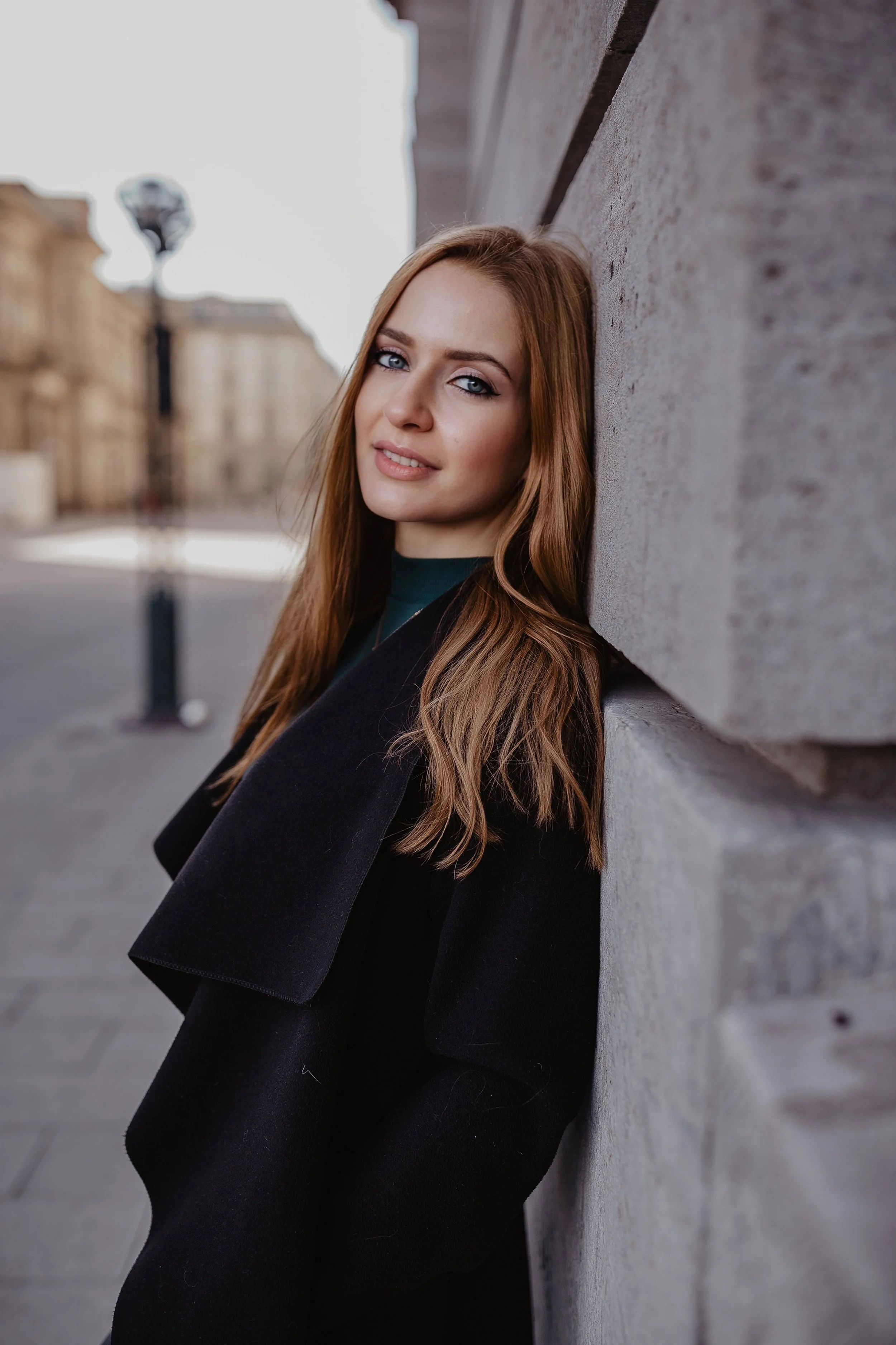 Junge Frau mit langen braunen Haaren lehnt an einer Steinwand im Freien.