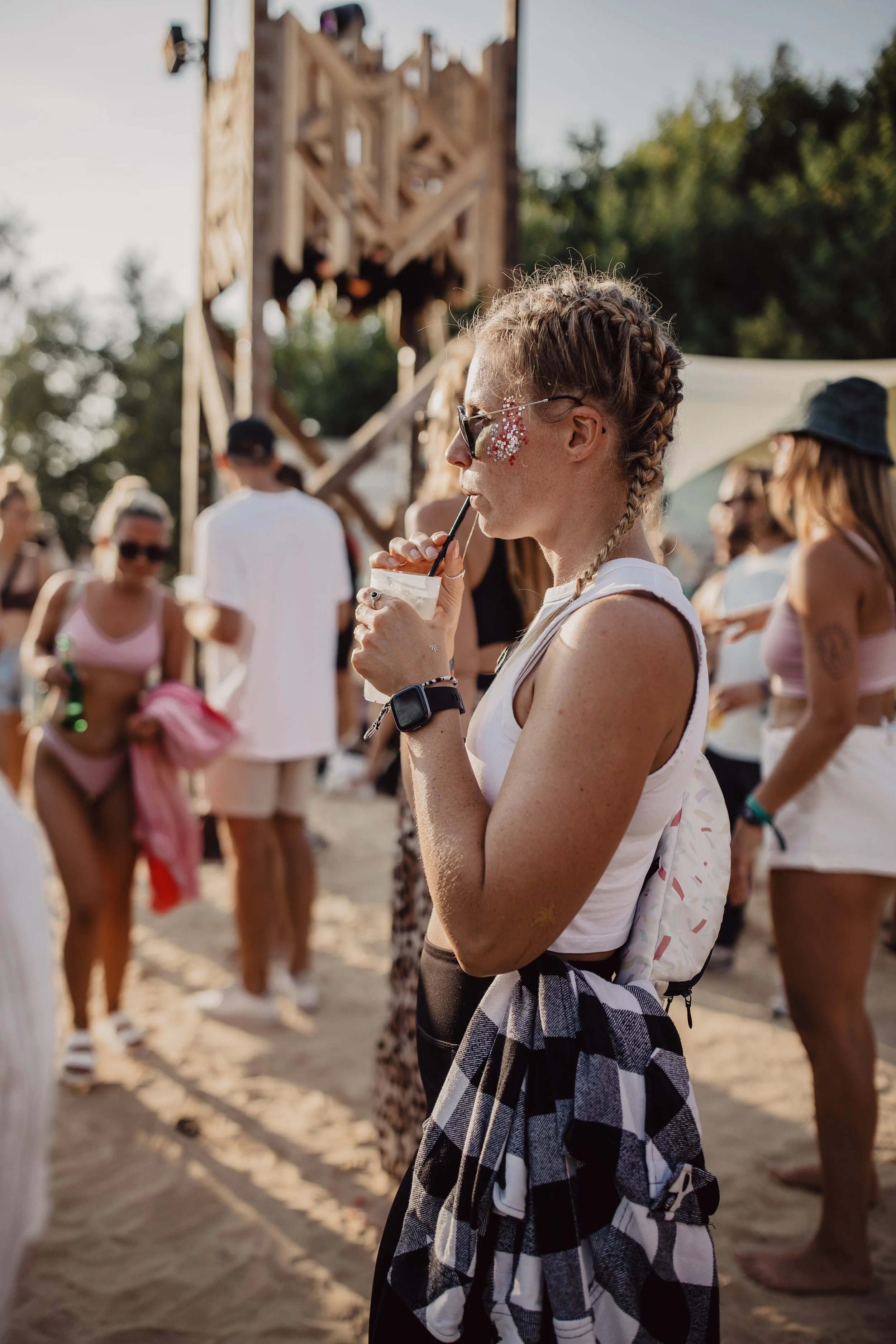 Junge Frau mit Flechtzopf und Sonnenbrille trinkt einen Drink bei einem Festival im Freien, umgeben von anderen Festivalbesuchern, umgeben von Bäumen und einer Holzkonstruktion im Hintergrund.