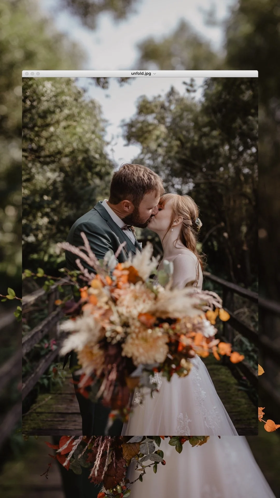Ein frisch verheiratetes Paar küsst sich auf einer Brücke, die von Bäumen umgeben ist, mit einem Blumenstrauß im Vordergrund.