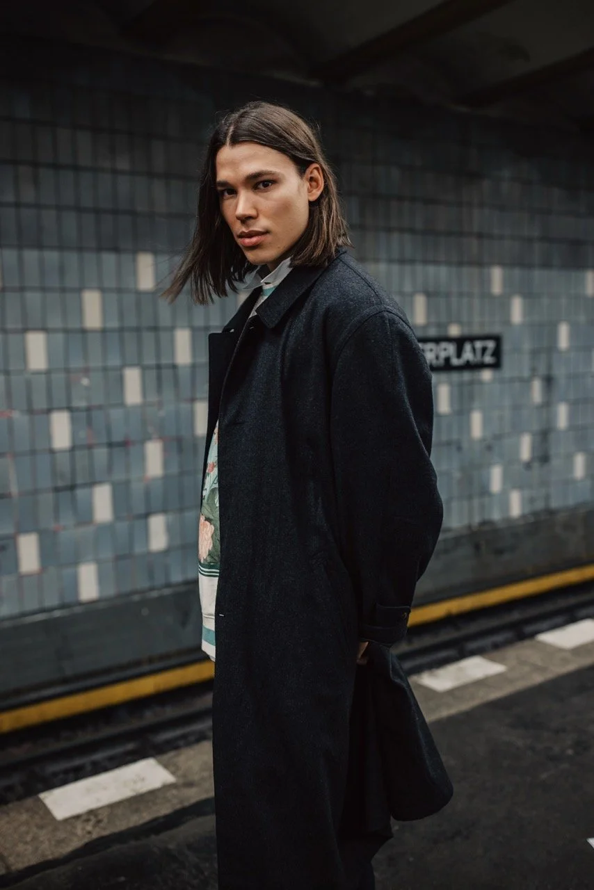 Junger Mann mit langem, glattem Haar steht in einer U-Bahn-Station. Er trägt einen schwarzen Mantel und schaut zur Kamera. Im Hintergrund eine Wand mit kleinen Kacheln und einem Schild mit der Aufschrift 'PLATZ'.