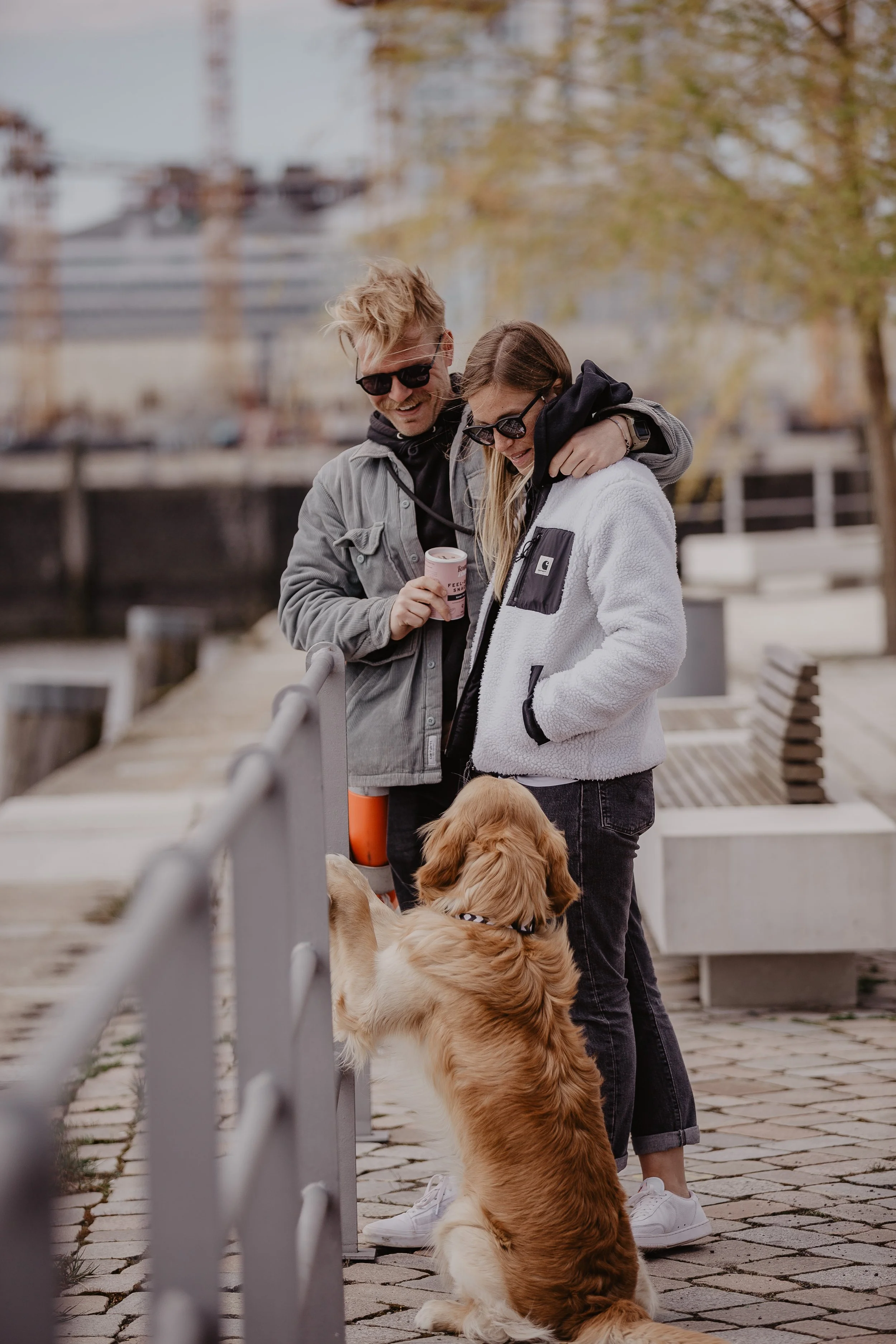 Ein lächelndes Paar mit einem Hund am Wasser entlang, im Hintergrund eine Brücke und Bäume im Herbst, die beiden tragen Jacken und Sonnenbrillen.