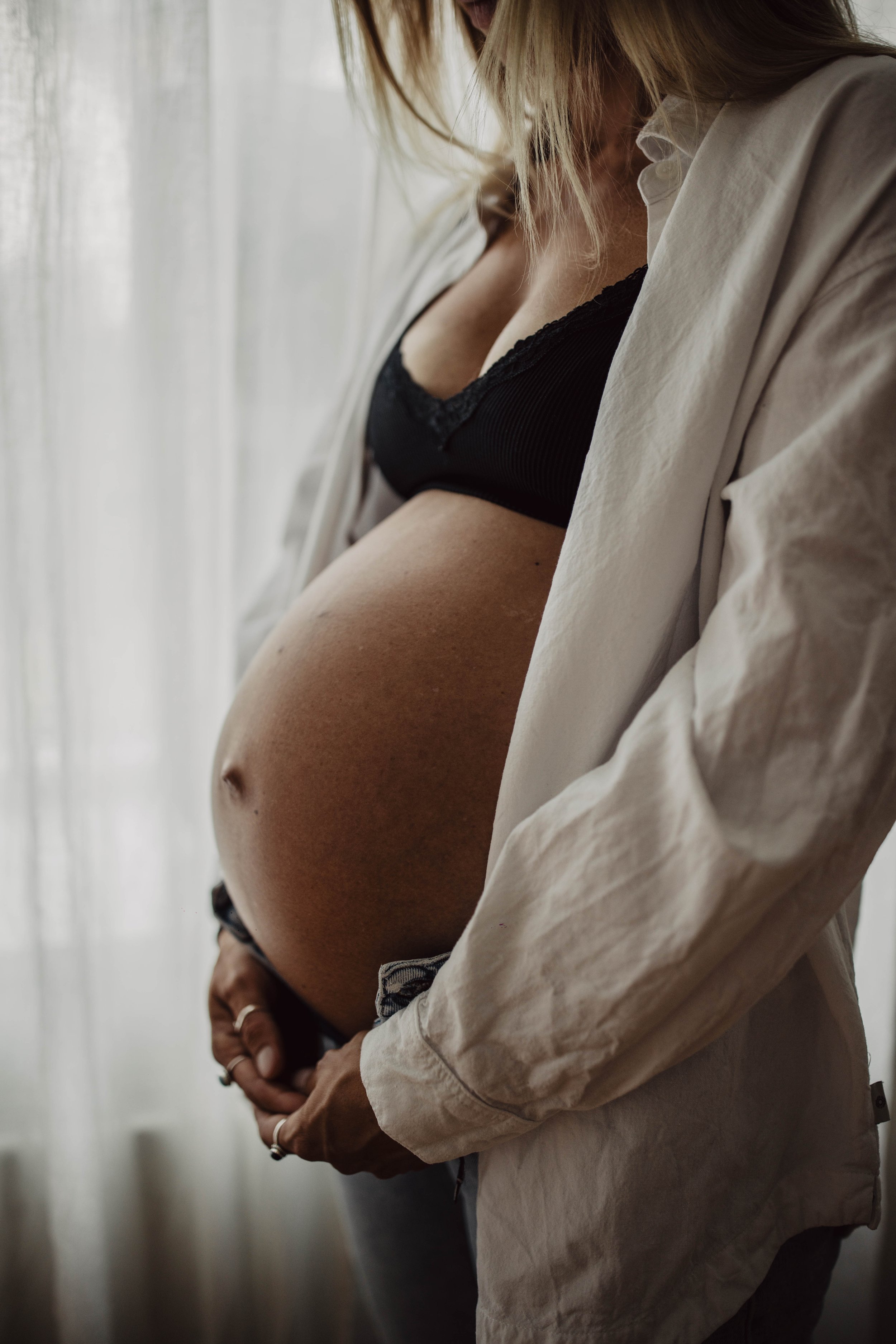 Schwangere Frau in dunklem Top und beige Jacke steht vor einem Fenster, mit ihren Händen auf dem Bauch.