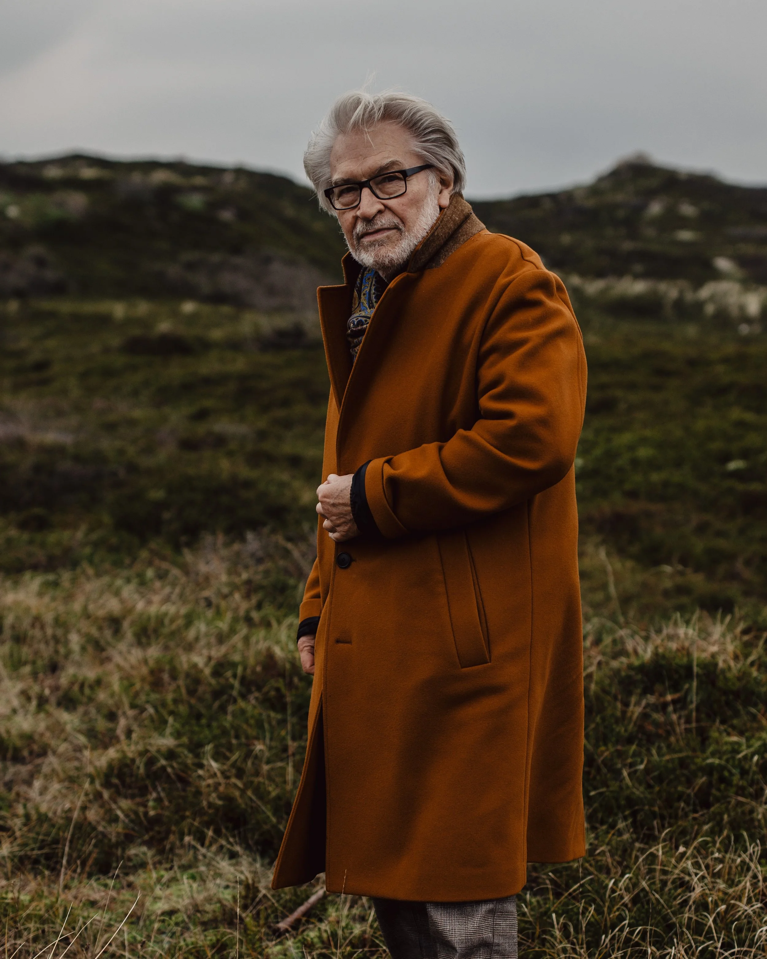 Älterer Mann mit grauem Haar und Brille steht in einem grasbewachsenen Gelände bei bewölktem Himmel, trägt einen braunen Mantel.